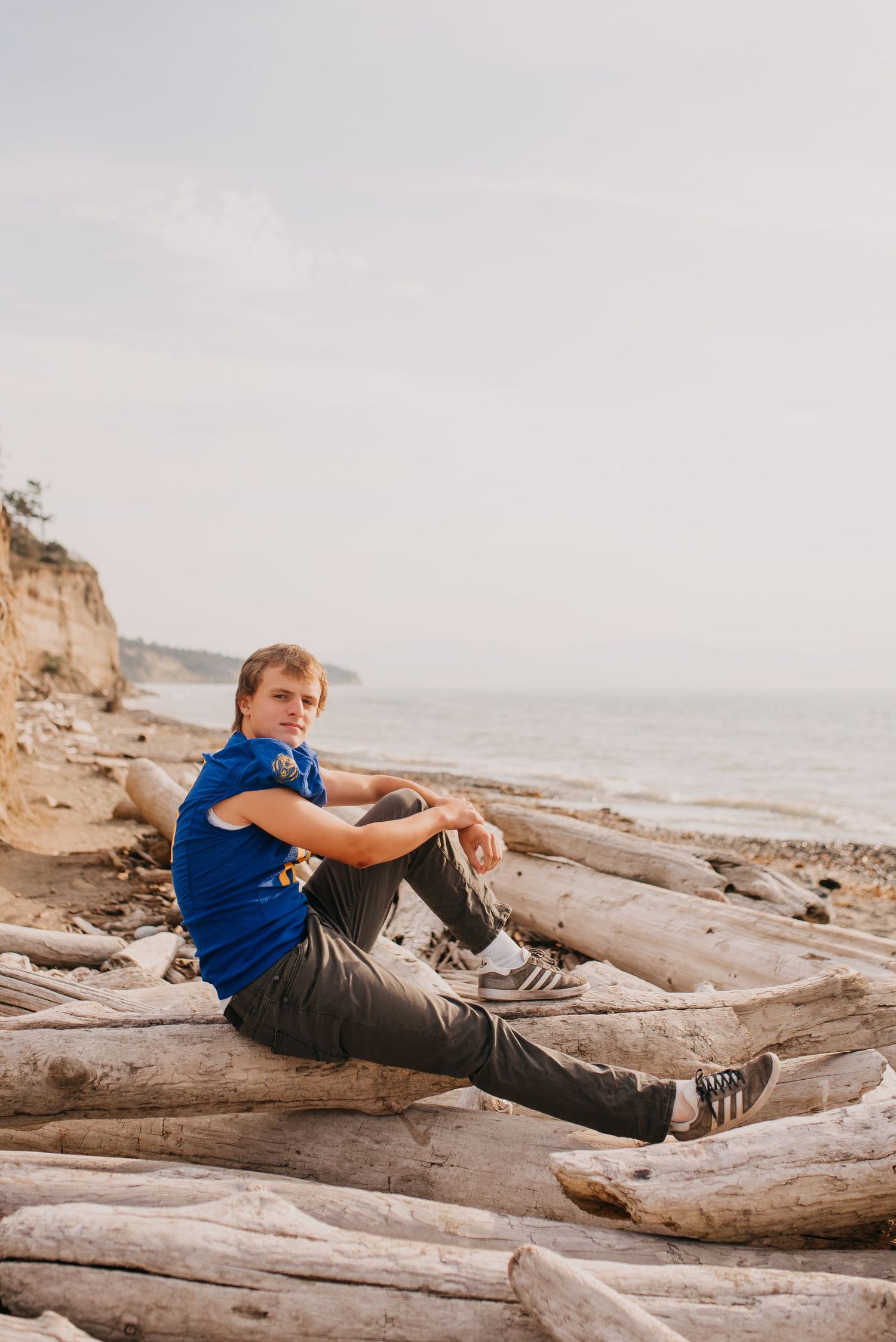 Teen boy in blue shirt sitting on driftwood logs on rocky beach for outdoor senior portraits.