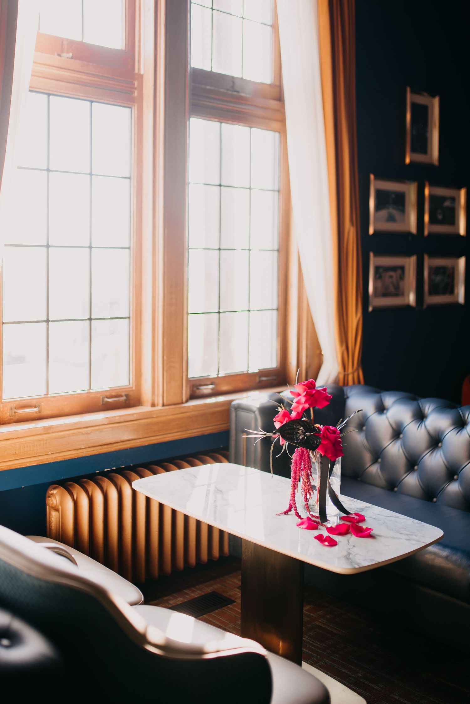 Elegant lounge interior with tufted sofa, bright windows, and pink floral arrangement on marble table.