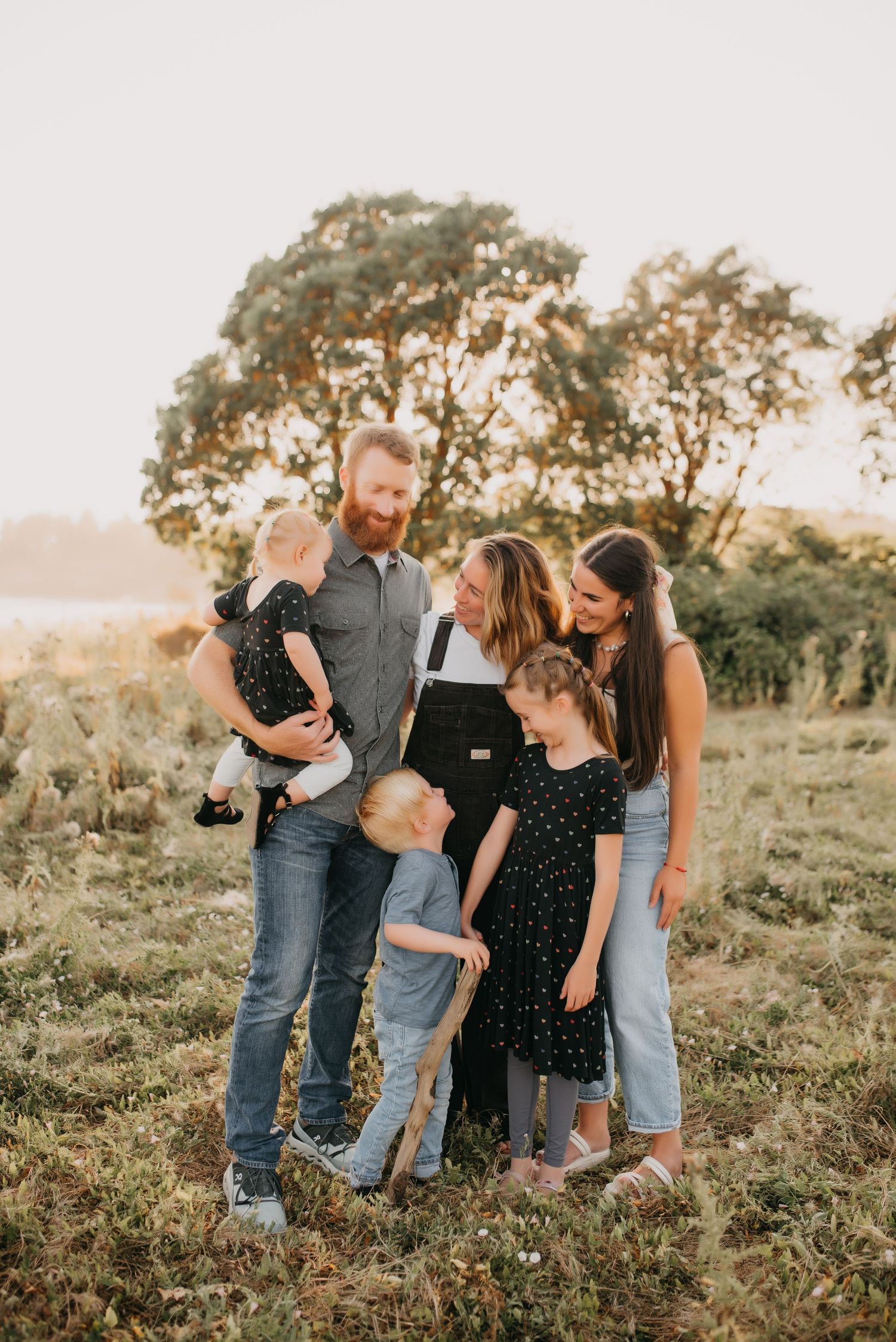 Large family of six gathered closely together smiling in sunlit meadow at golden hour.