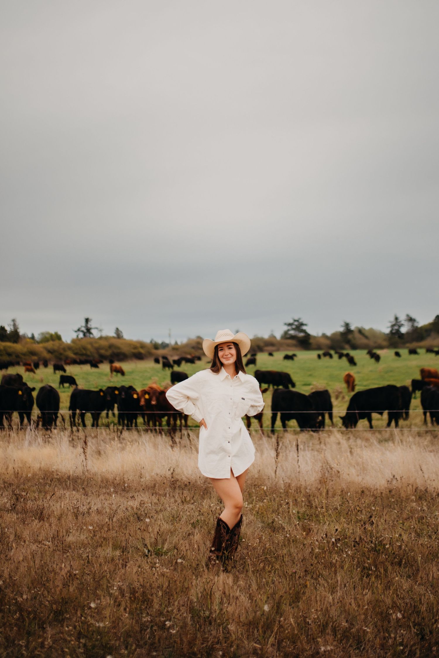 Young woman in white dress and cowboy hat posing in field with black cattle grazing behind.