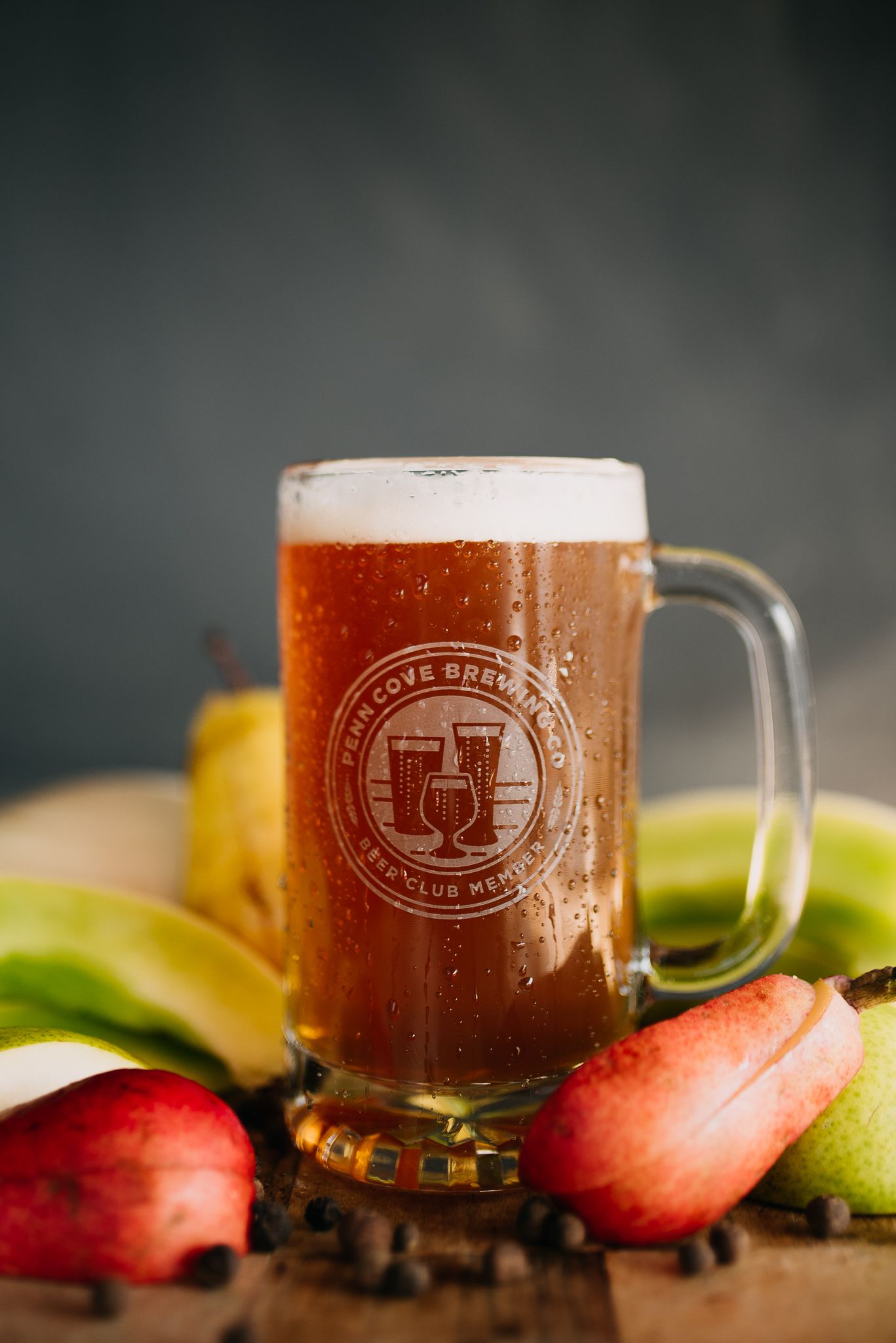 Branded craft beer mug surrounded by fresh pears and apples on a wooden surface.