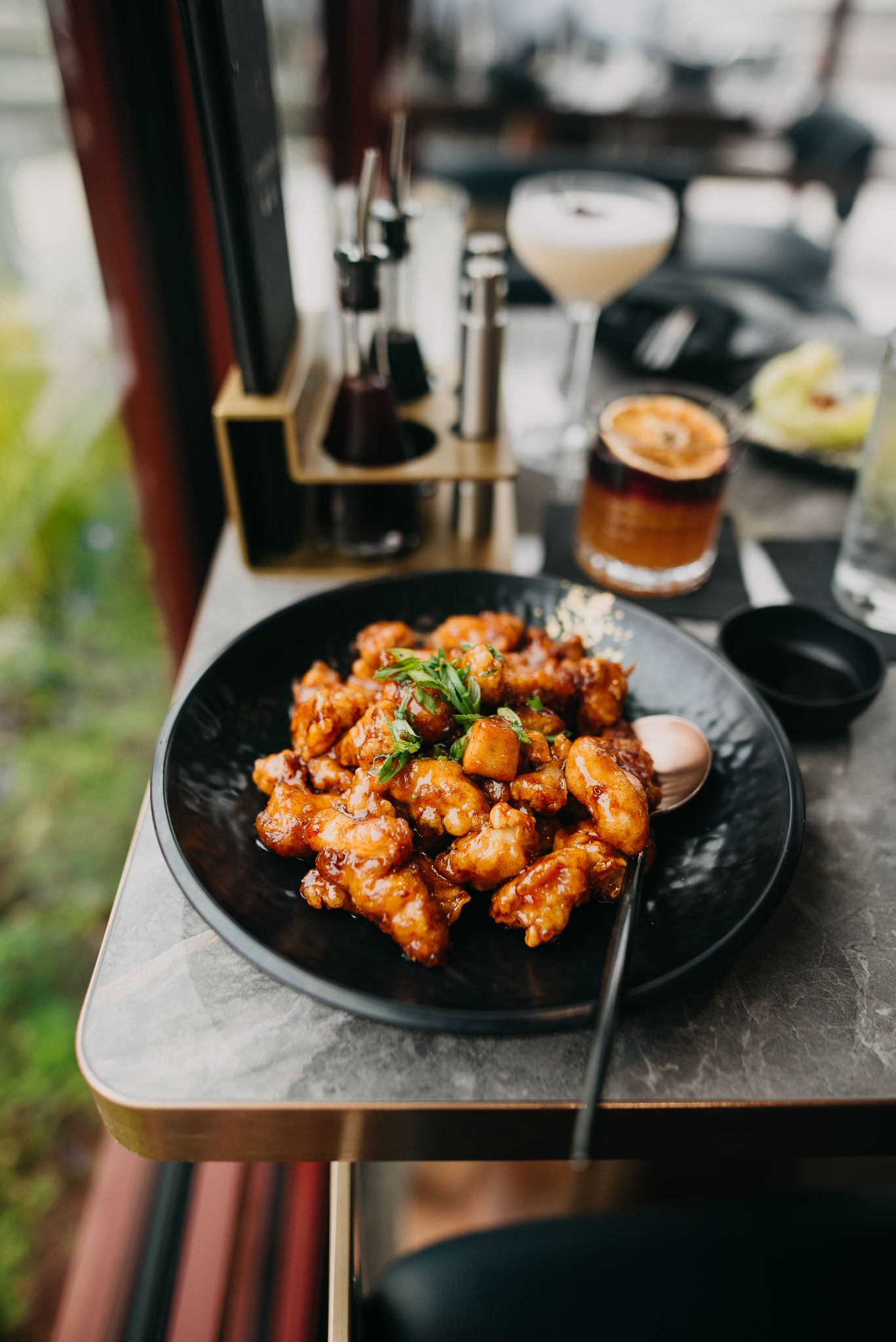Glazed Korean fried chicken on a black plate served with cocktails at a restaurant table.