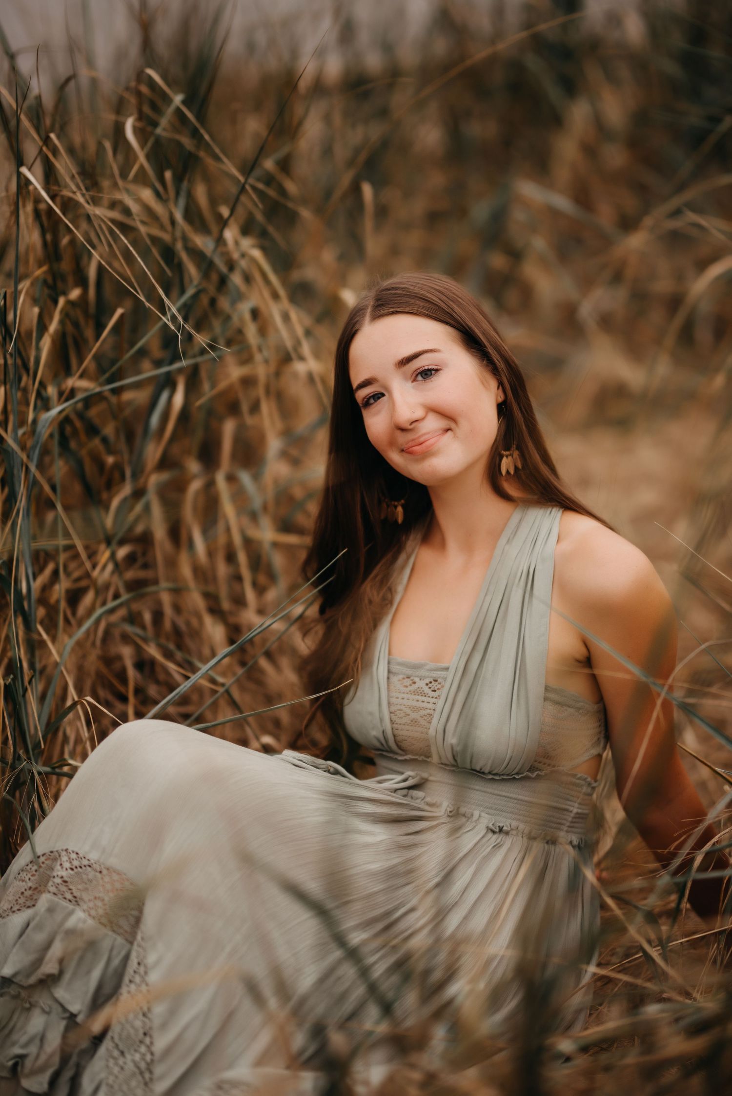 Young woman in sage green boho dress sitting in golden tall grass field, smiling naturally.