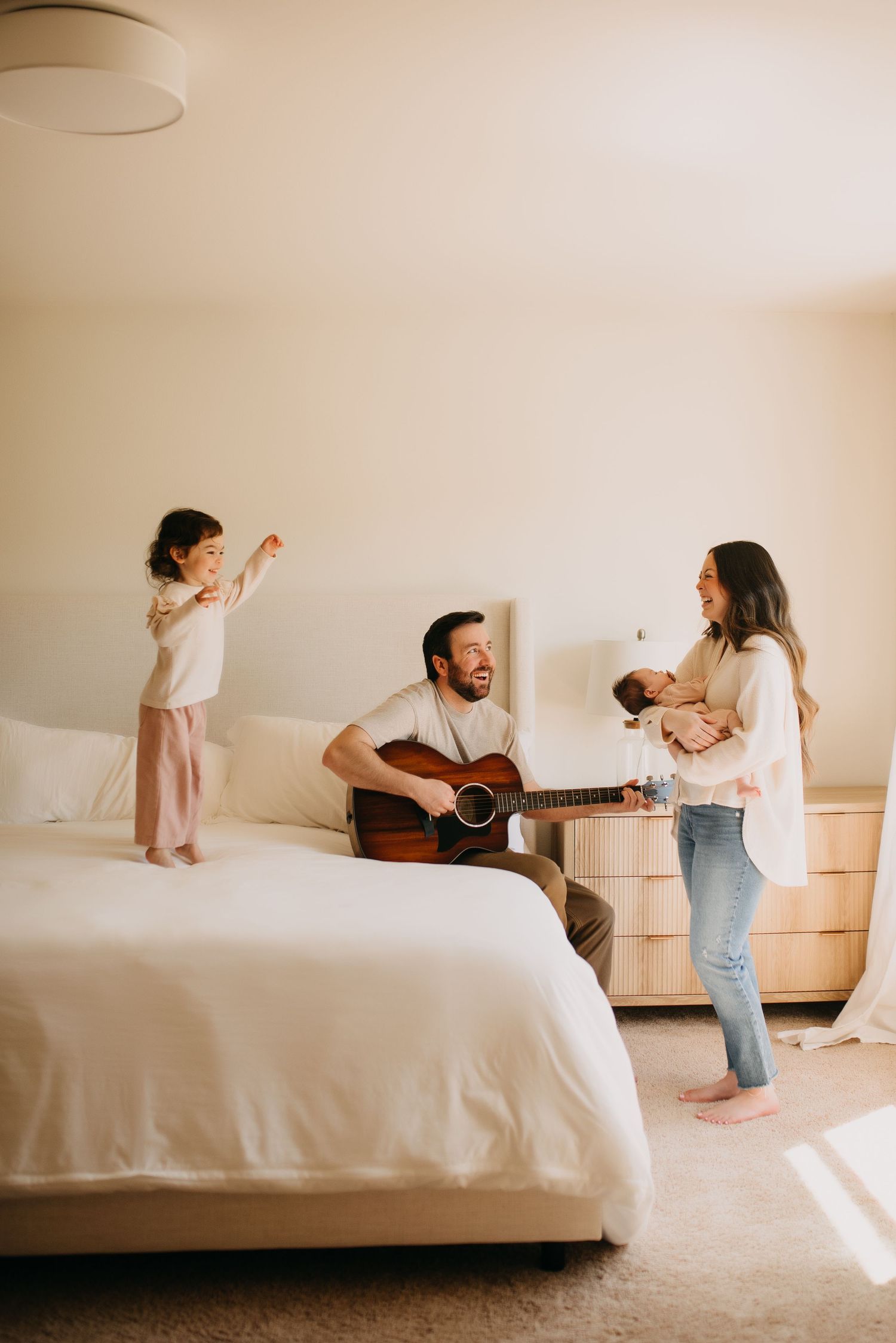 Father playing acoustic guitar on bed while daughter dances and mother holds newborn baby.