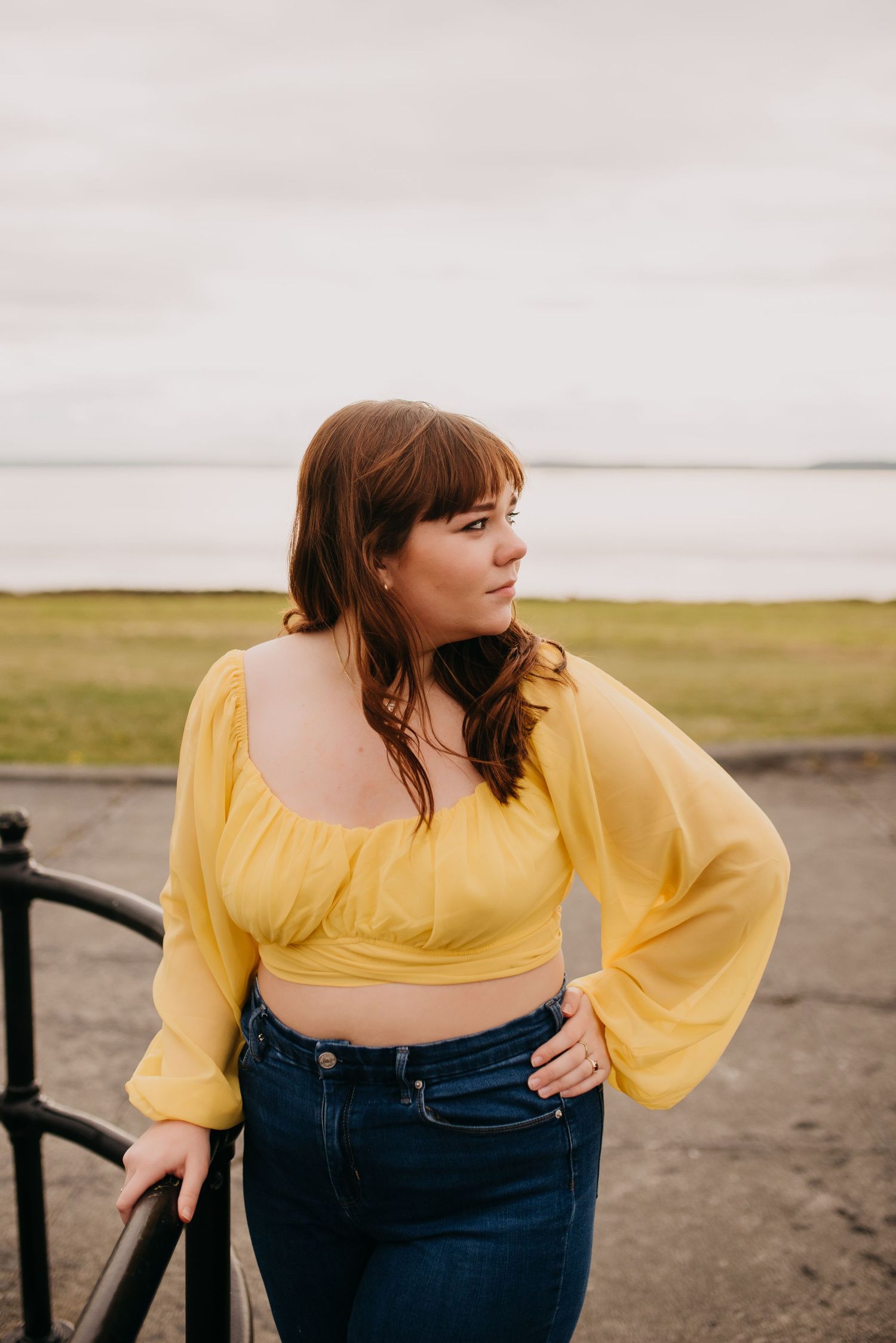 Young woman in yellow crop top and jeans posing by iron railing with ocean view in background.