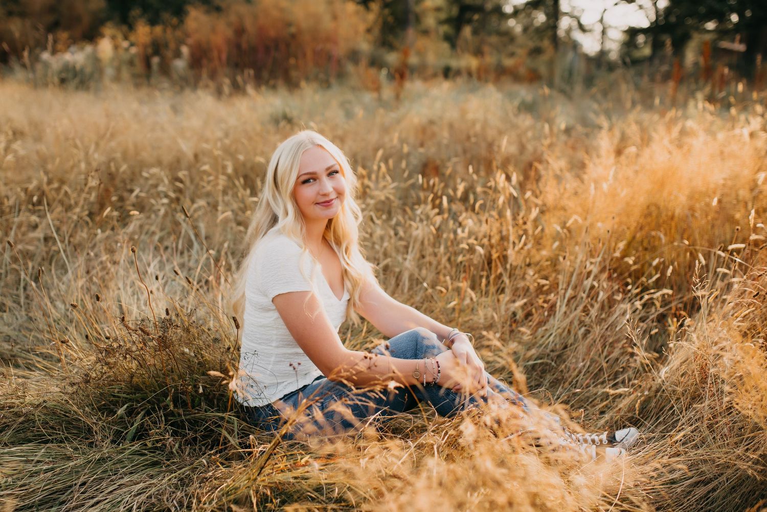 Smiling blonde high school senior sitting in golden grass field wearing white tee and jeans.