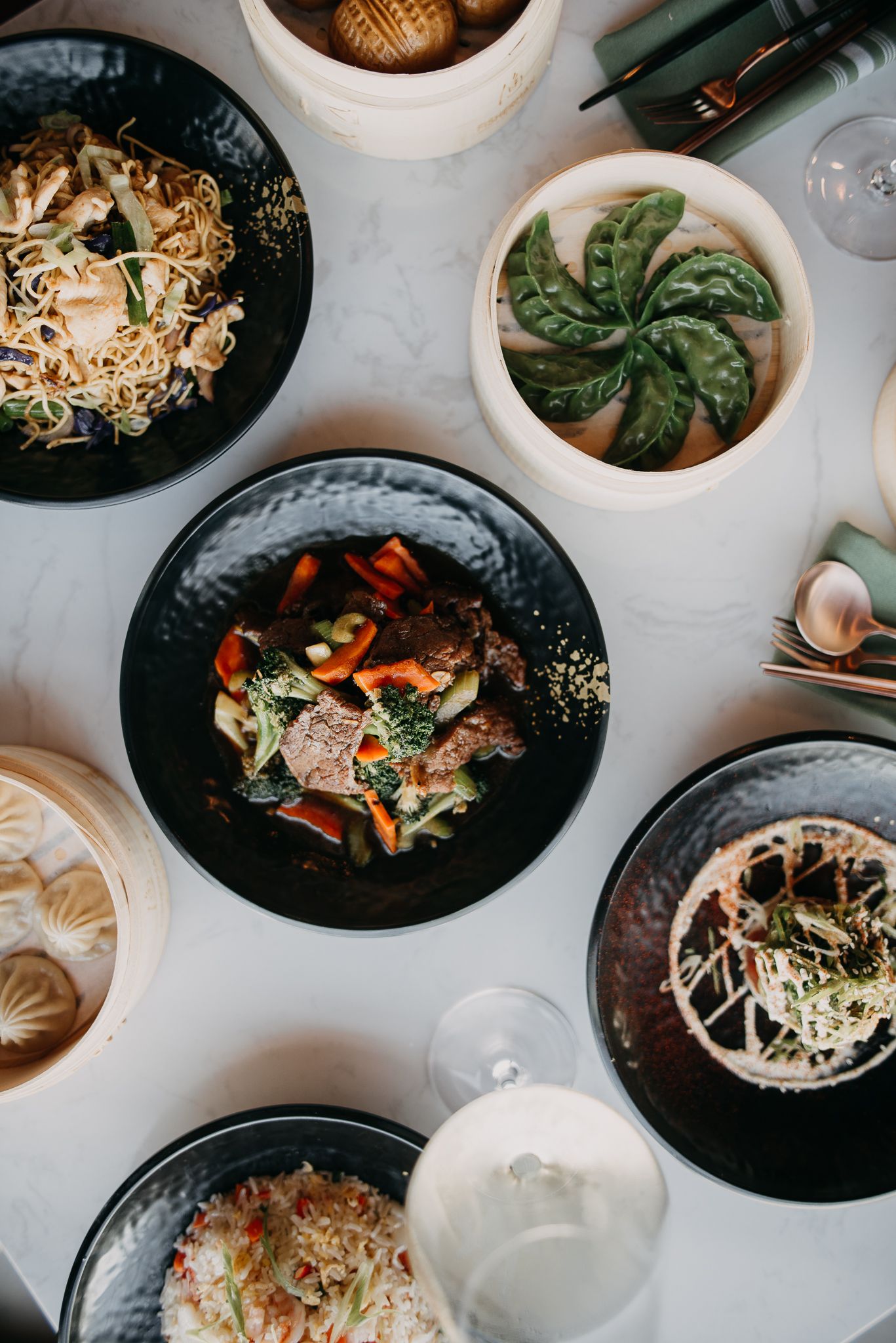 Aerial view of Asian feast featuring stir-fry, noodles, dumplings and steamed dishes on marble.