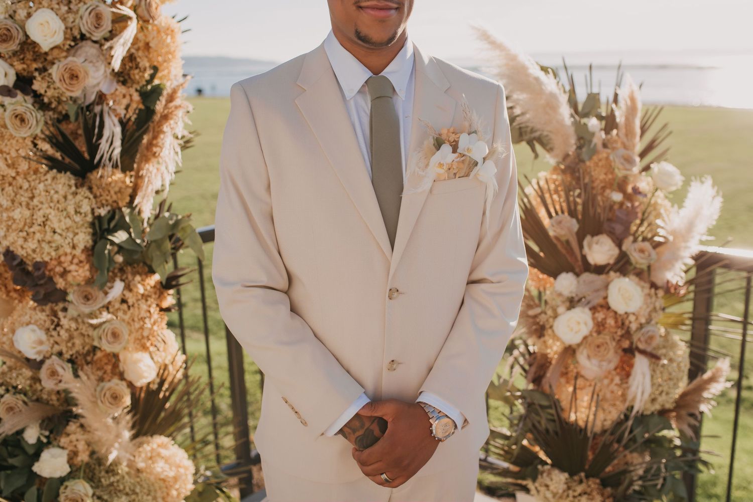 Groom in beige suit stands at floral altar adorned with dried pampas grass and white blooms.