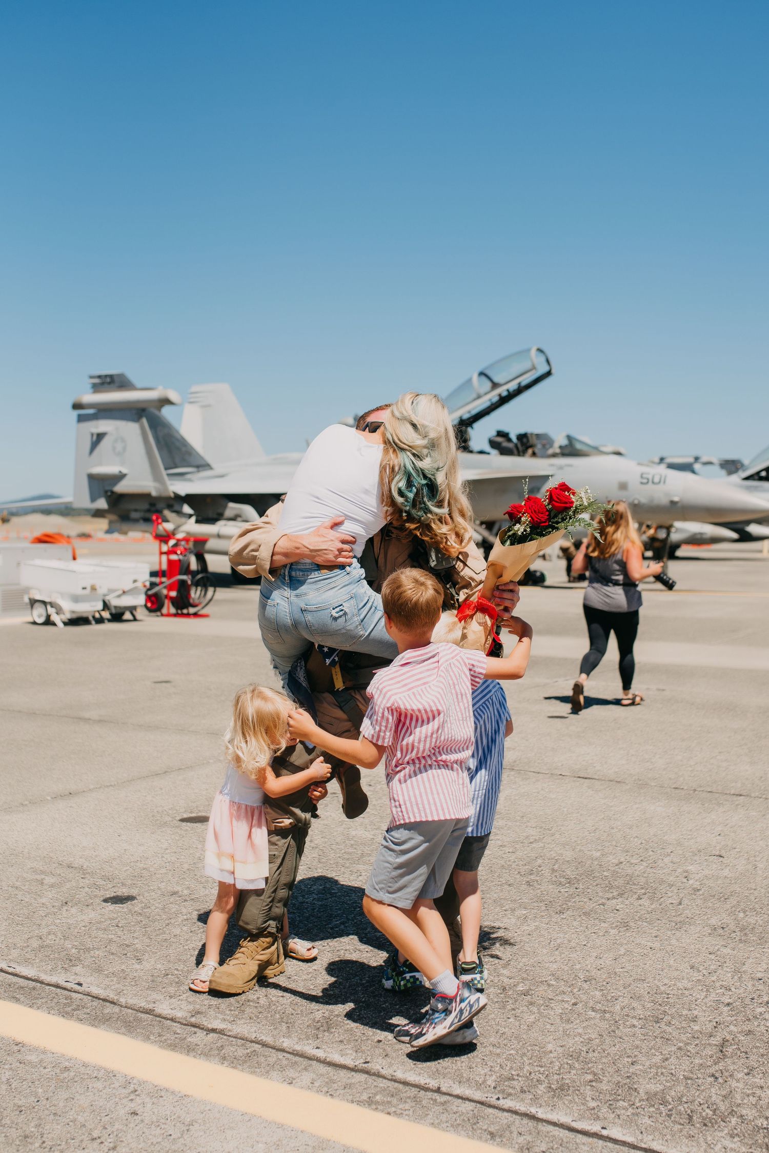 Military family reunion on airfield tarmac with children hugging parent near fighter jet.