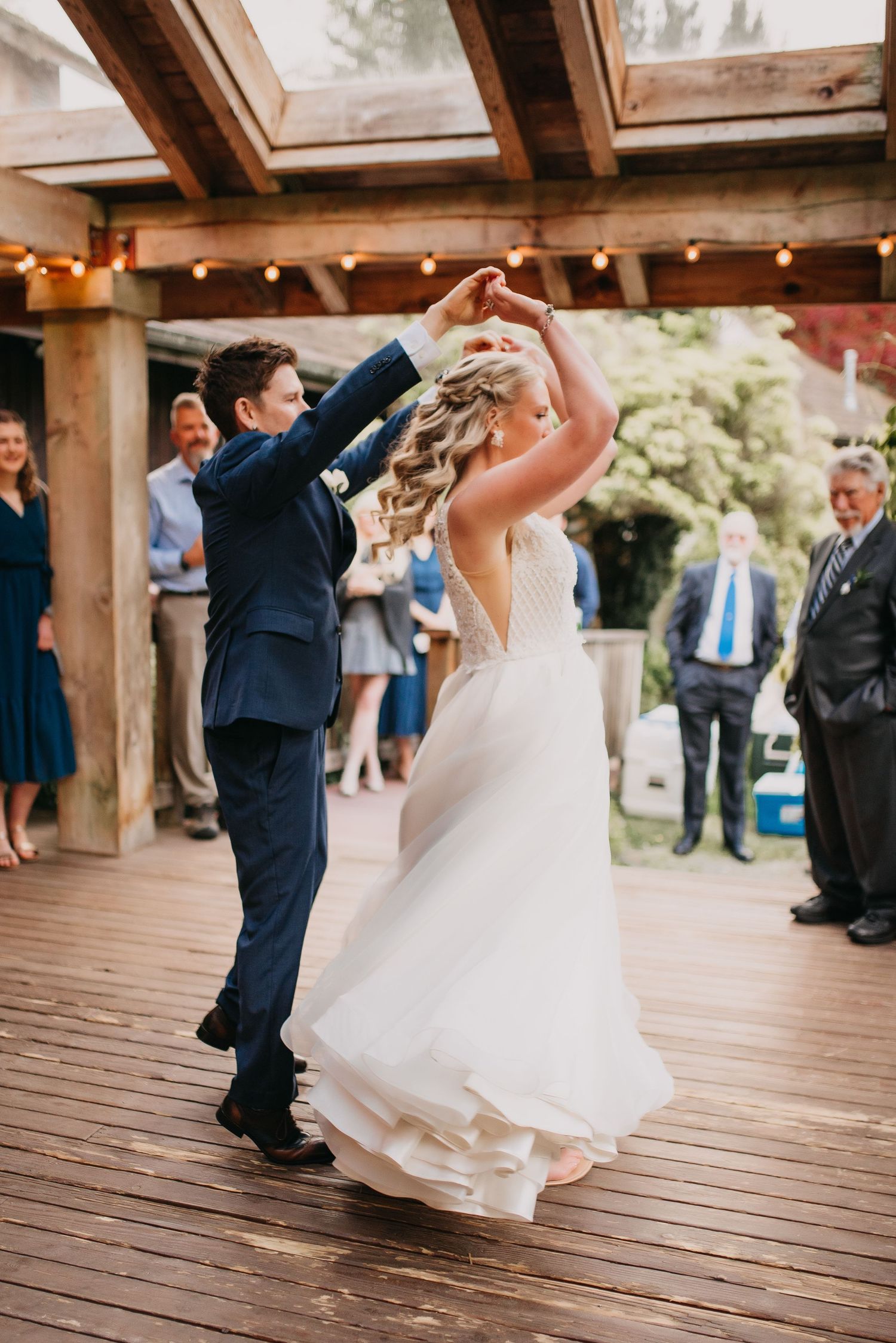 Groom in navy suit twirls bride in white lace gown on a rustic wooden deck with string lights.