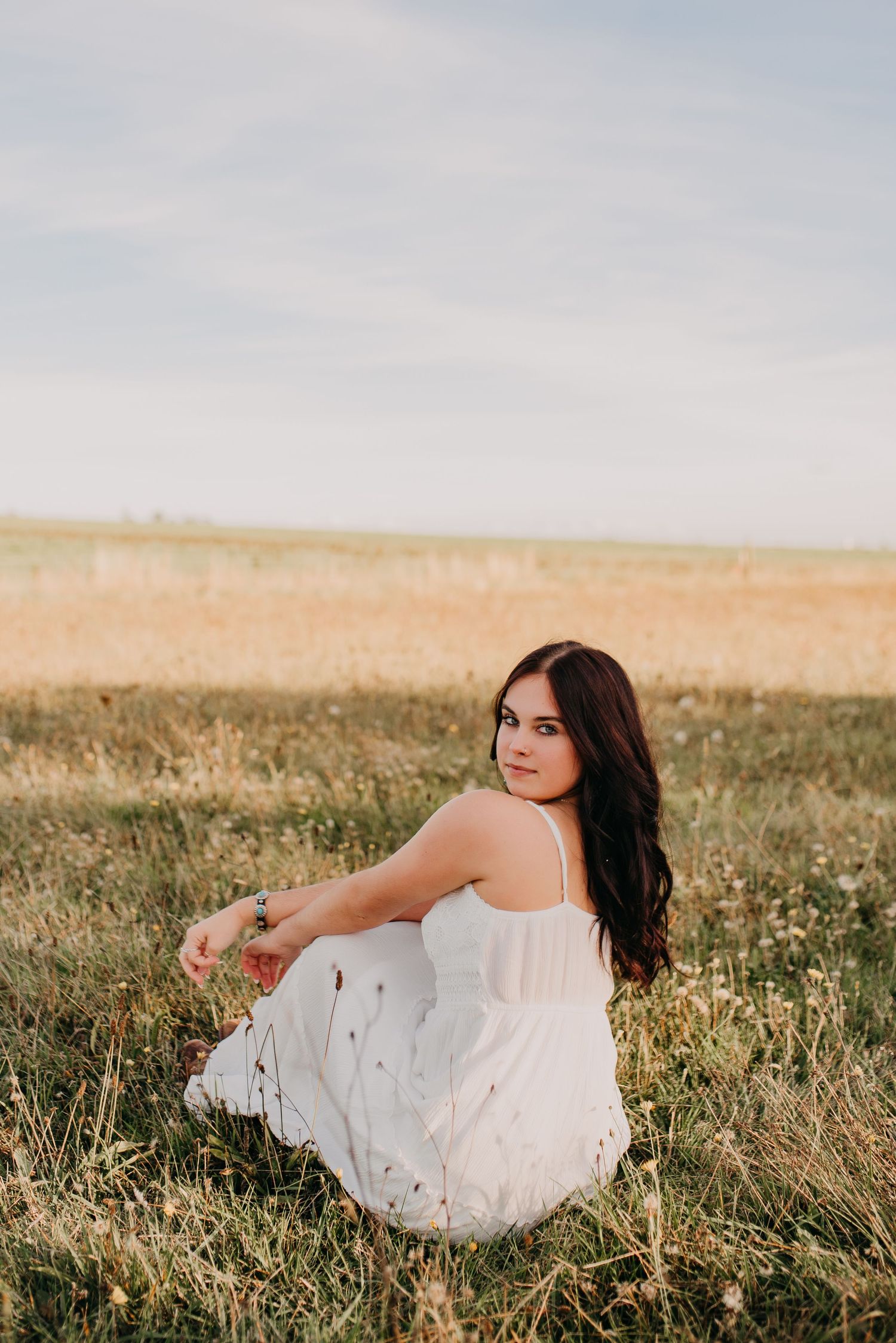 Young woman in white sundress sits gracefully in a sunlit meadow with golden fields in background.