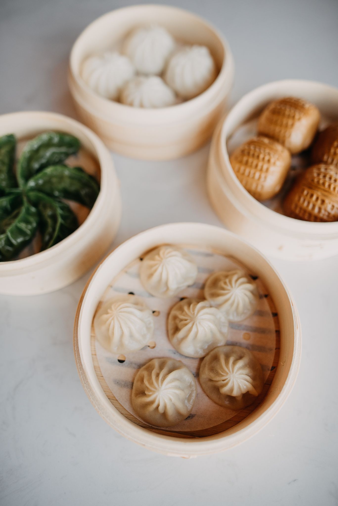 Four bamboo steamers filled with xiaolongbao, green dumplings and baked buns on white surface.