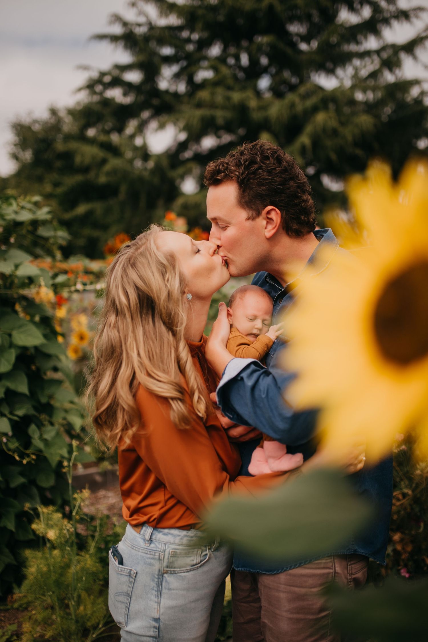 Couple kissing in a sunflower field while holding their newborn baby.