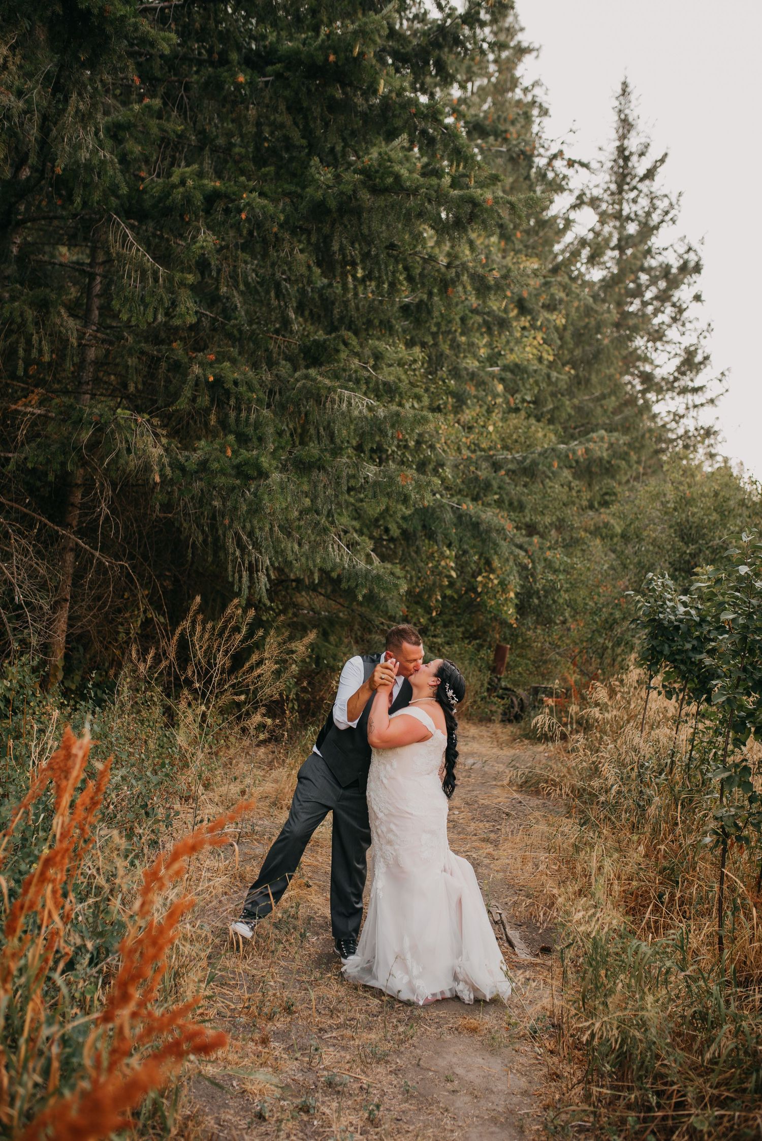 Bride and groom share a romantic kiss on a forest trail surrounded by tall evergreen trees and golden grass.