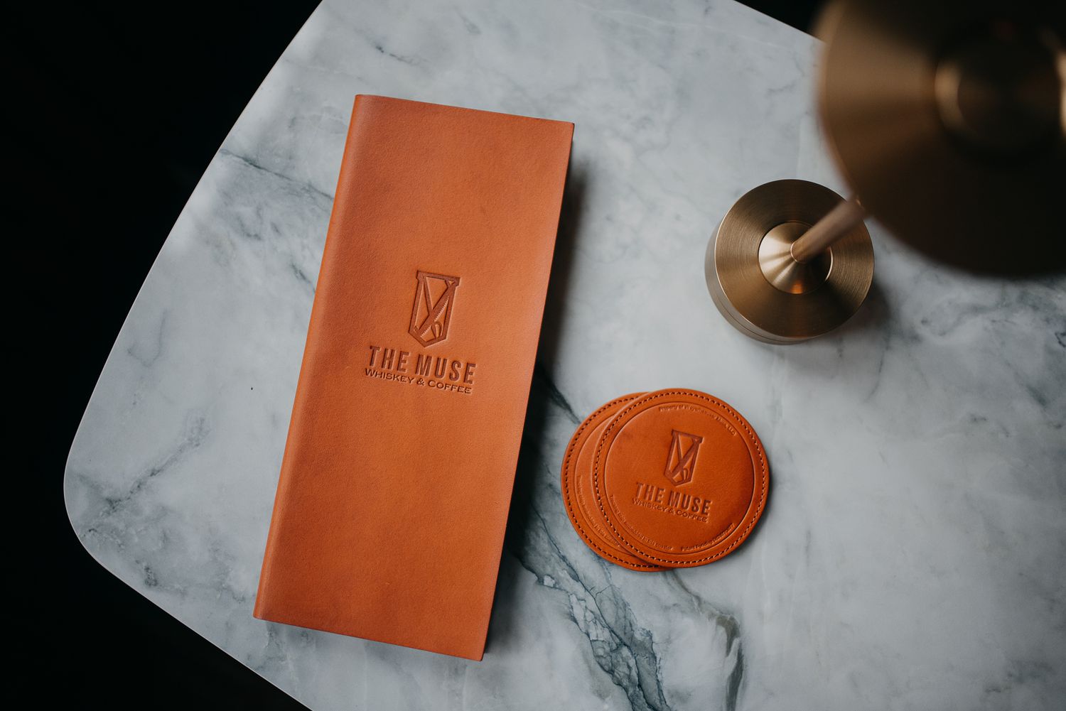 An elegant tan leather menu cover embossed with The Muse logo beside a wax seal on marble surface.
