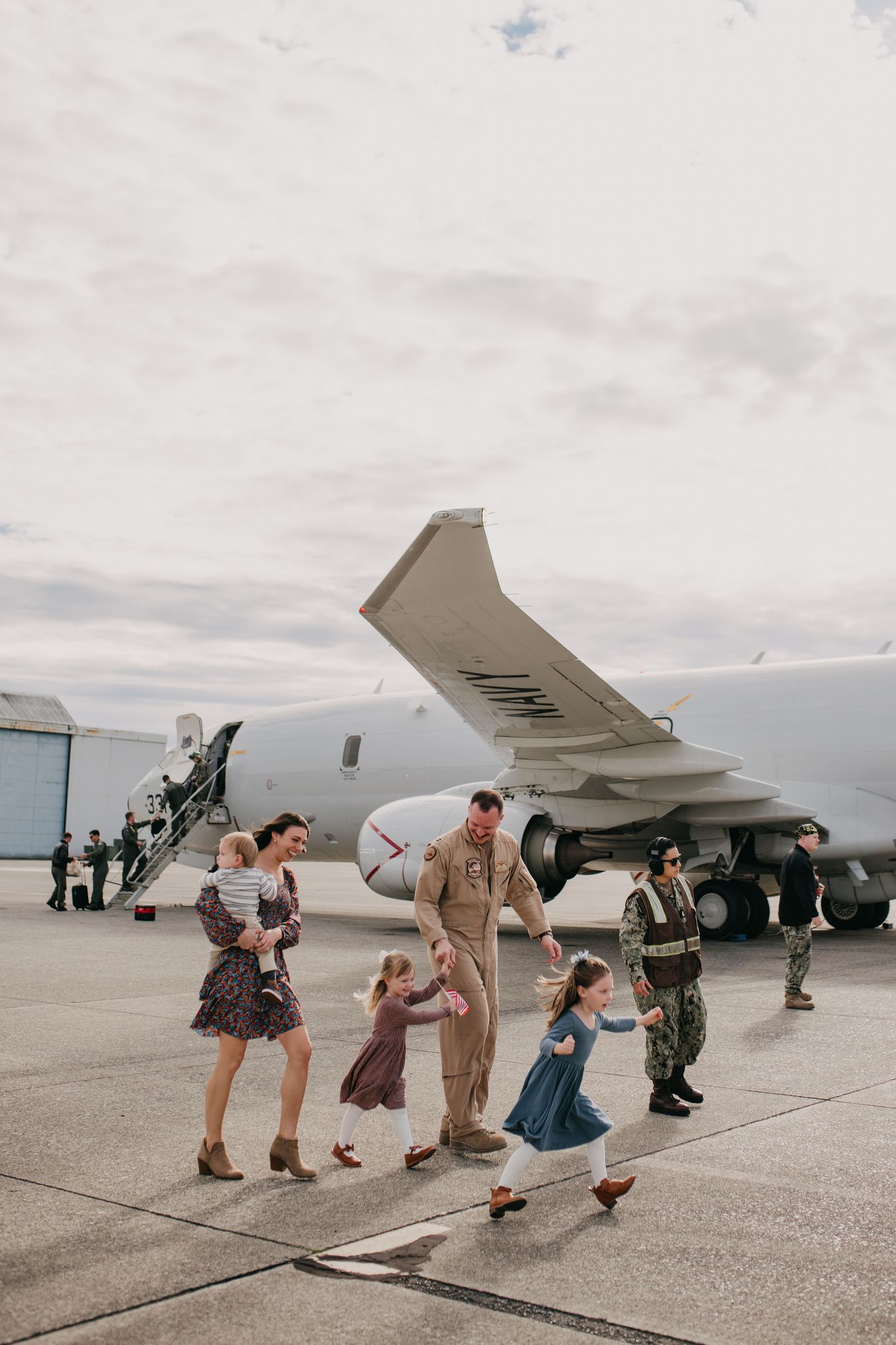 Military family reuniting on airport tarmac with young children beside large white aircraft.