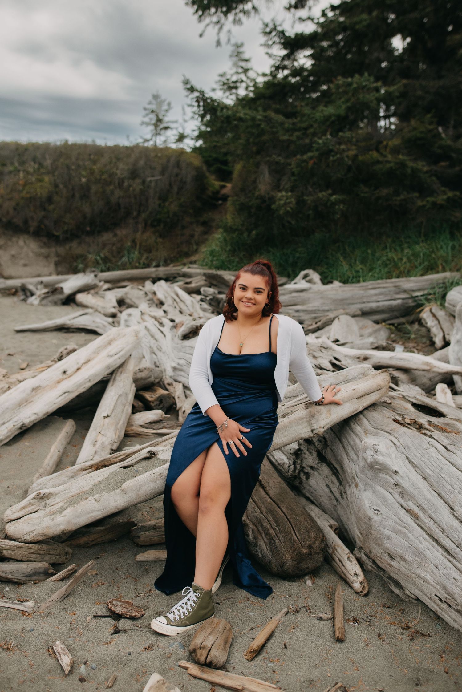 Senior photo session with a smiling young woman in blue dress and white cardigan posing on driftwood pile at Whidbey Island beach.