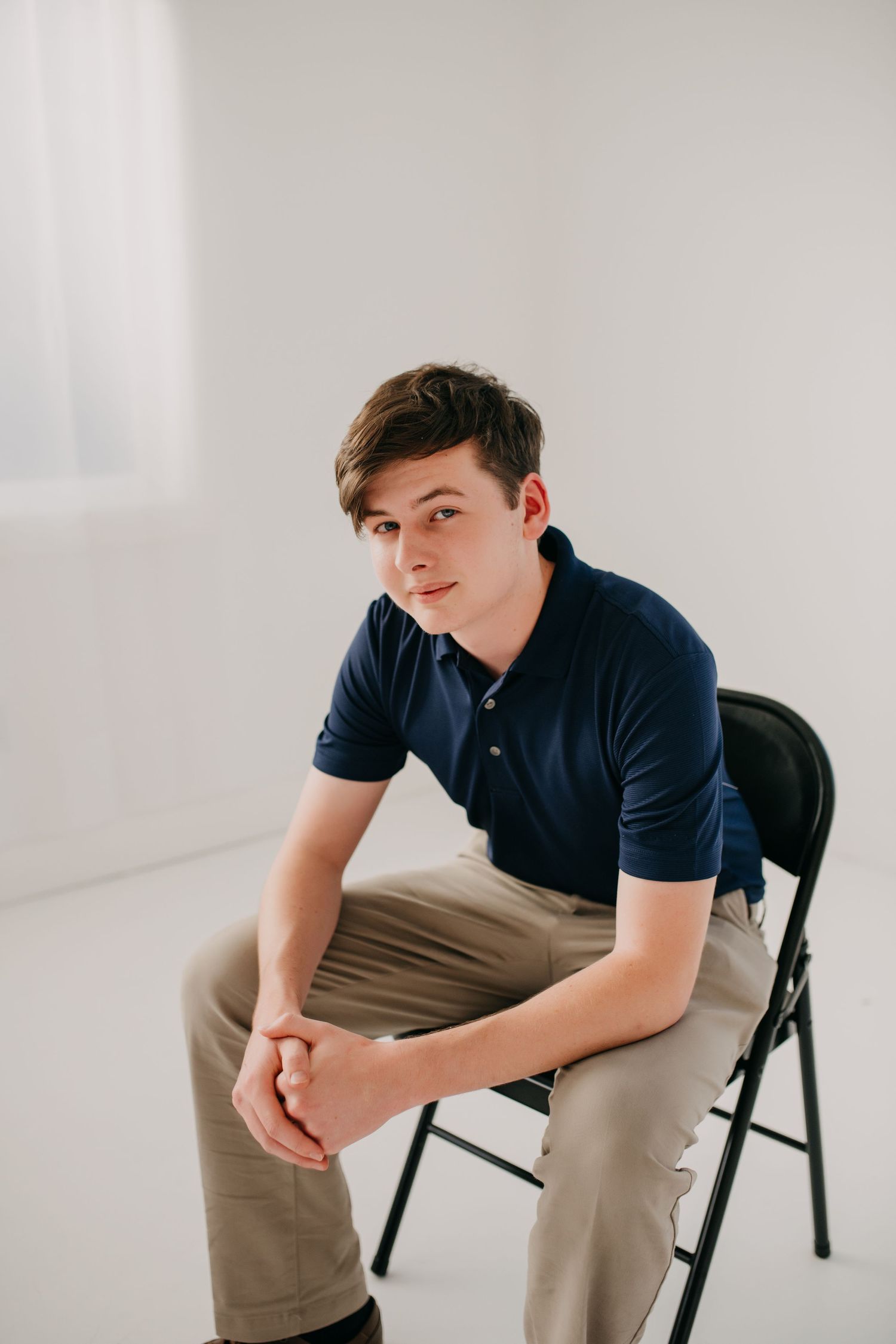 Young man in navy polo shirt sitting on a folding chair in a bright white studio, hands clasped.