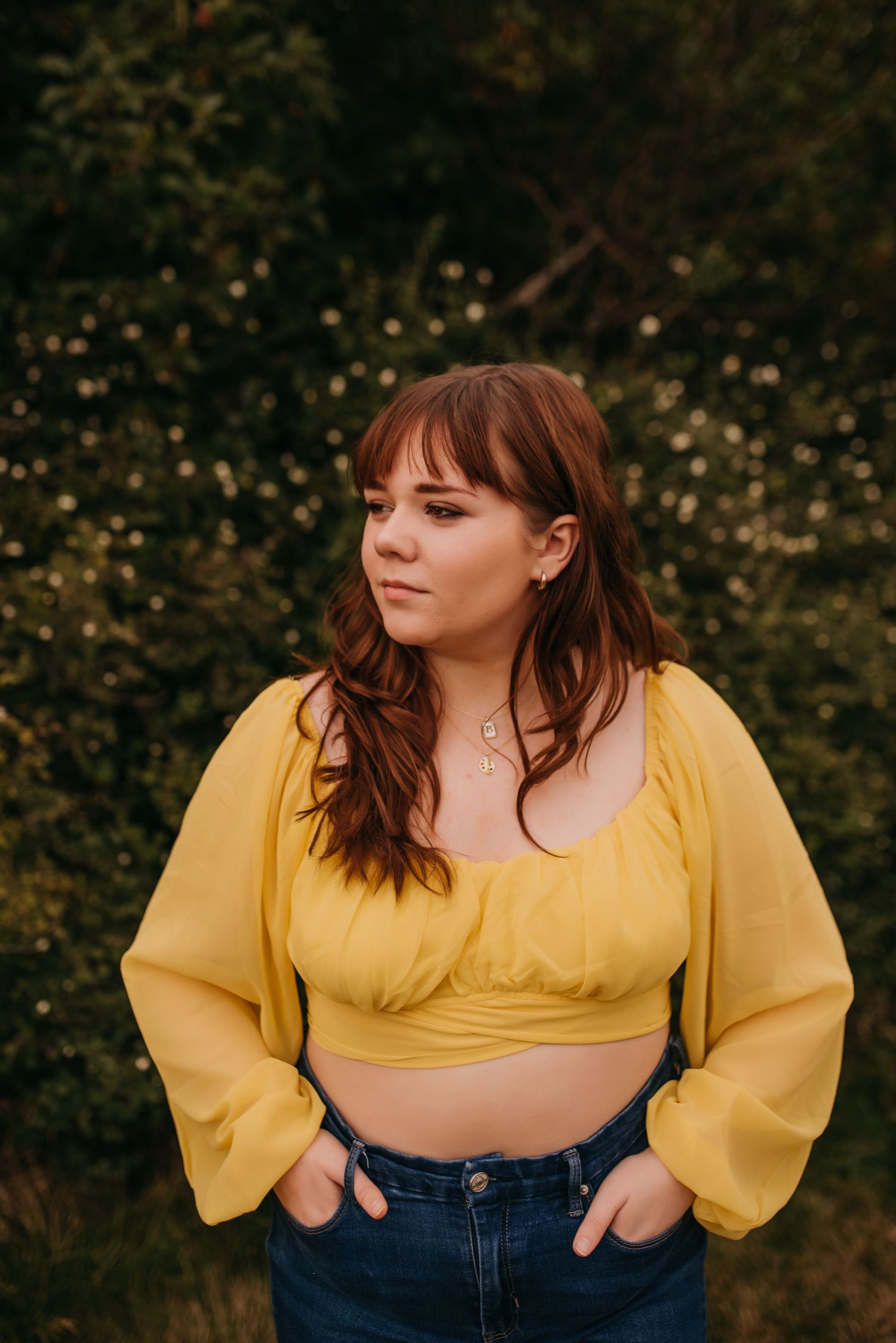Young woman in yellow crop top and jeans poses outdoors in front of green bushes.