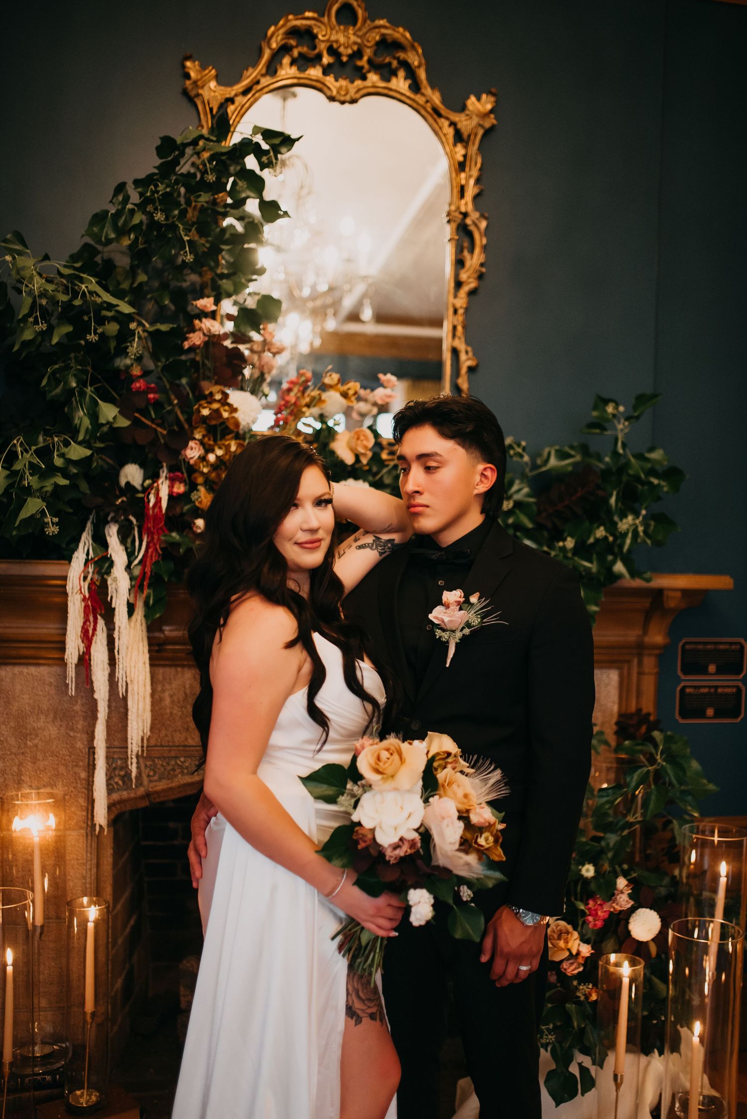 Bride and groom pose elegantly by a candlelit fireplace adorned with lush floral arrangements and greenery.