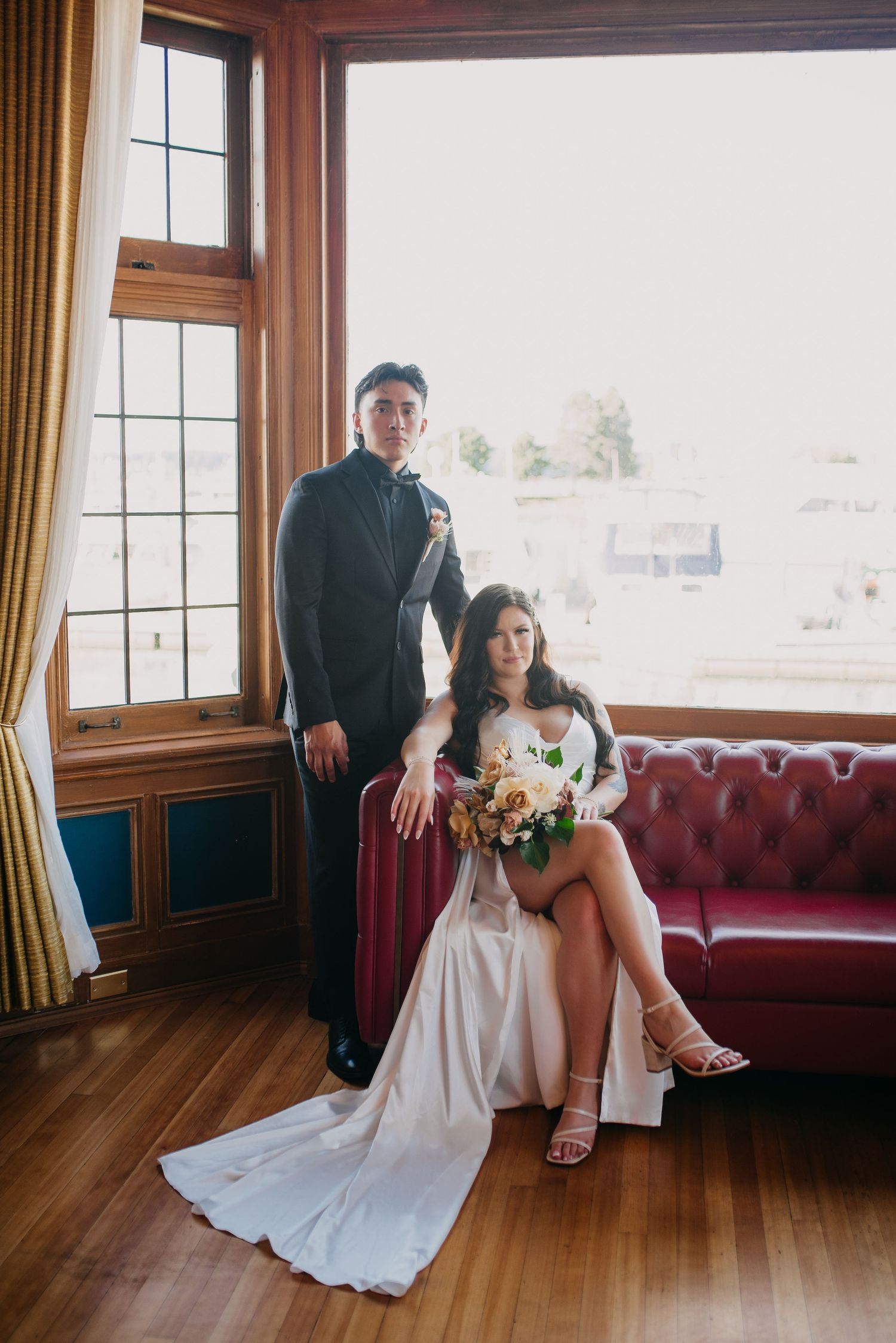 Bride and groom posing by a sunlit window on a red velvet sofa in a vintage-style venue.
