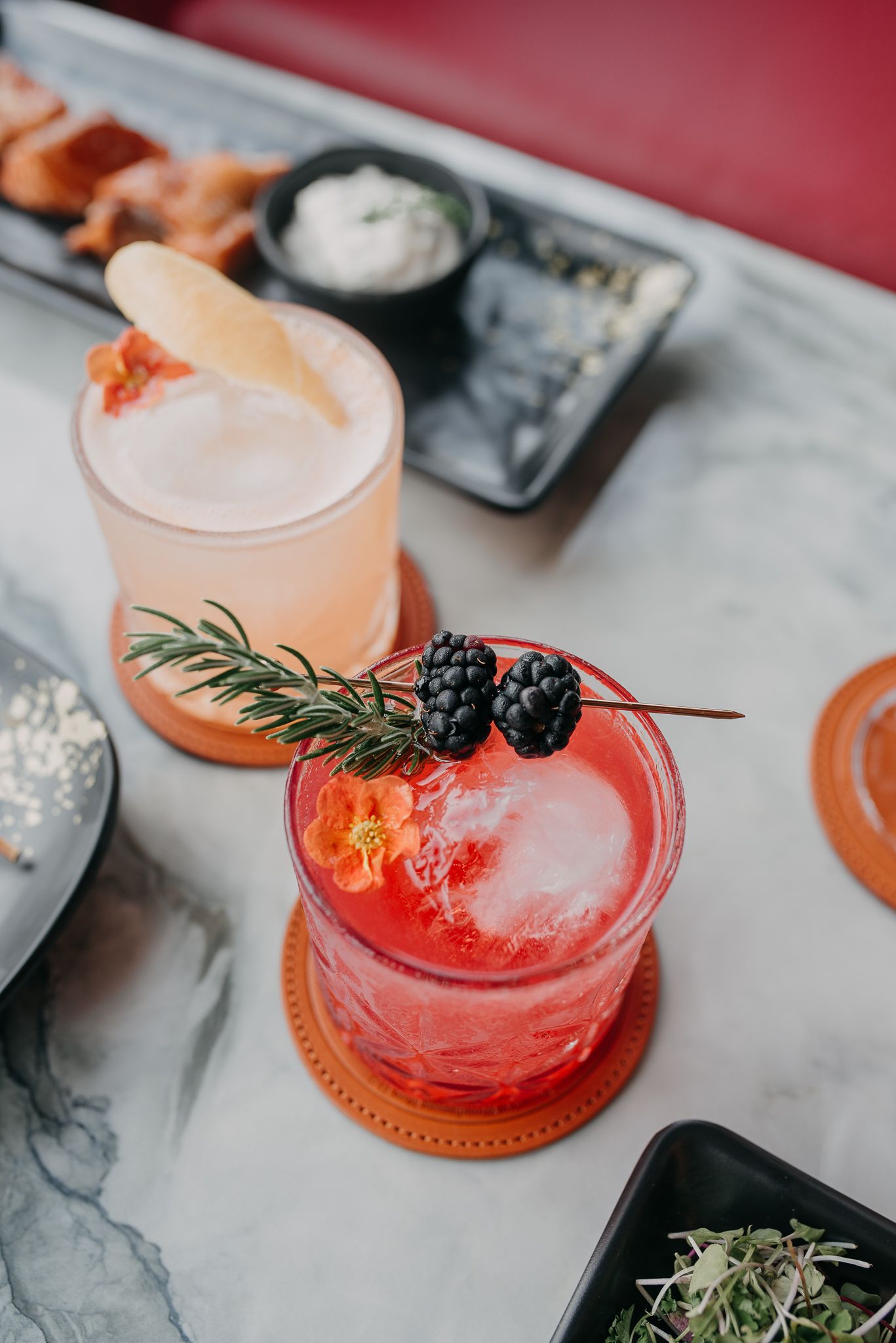 Two craft cocktails on marble bar, red drink garnished with blackberries and rosemary, pink drink with wafer garnish.