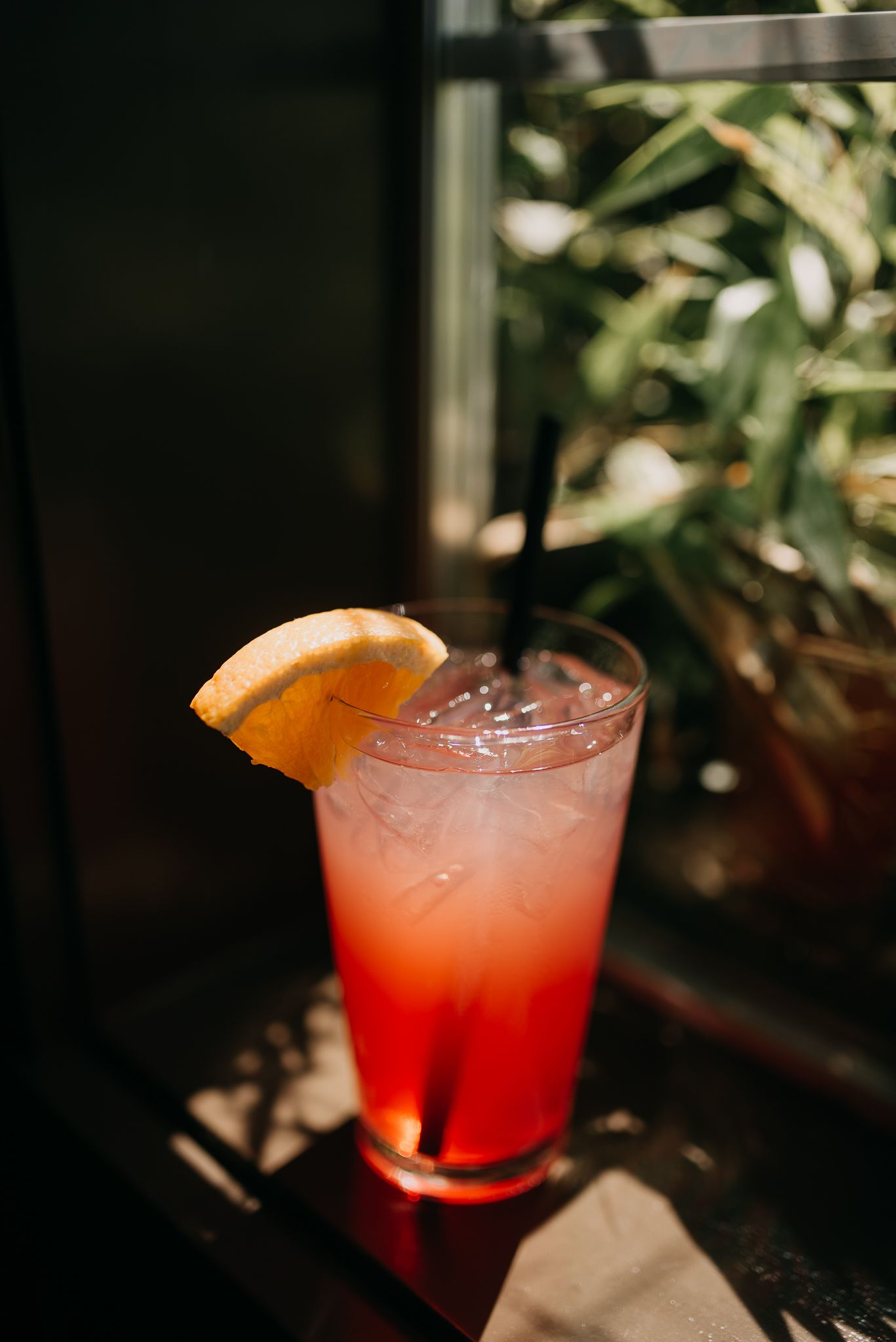 A vibrant red cocktail with ice, orange slice garnish, and black straw in a tall glass by a window.