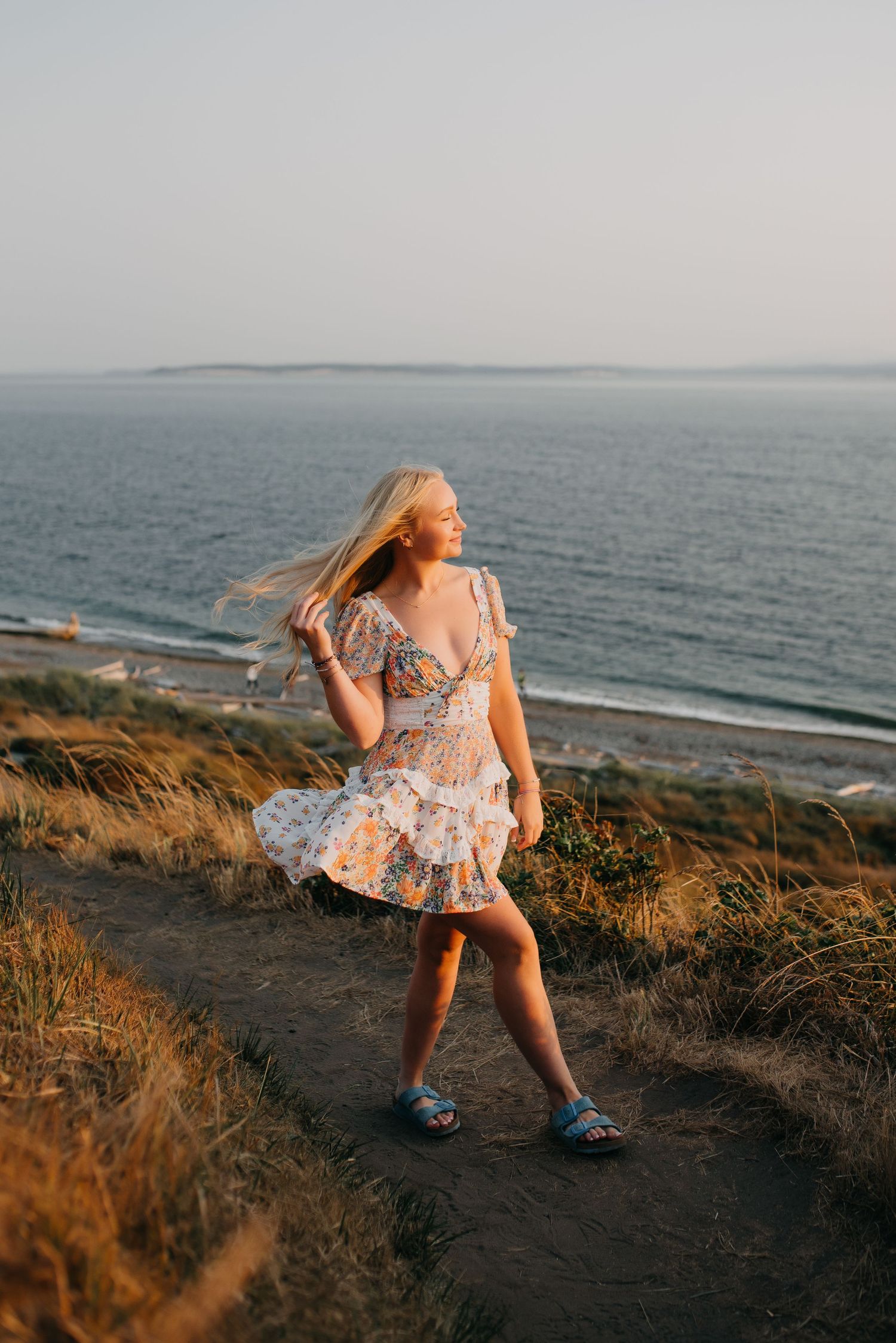Blonde woman in floral dress stands on a coastal bluff with wind-blown hair and ocean views at sunset.