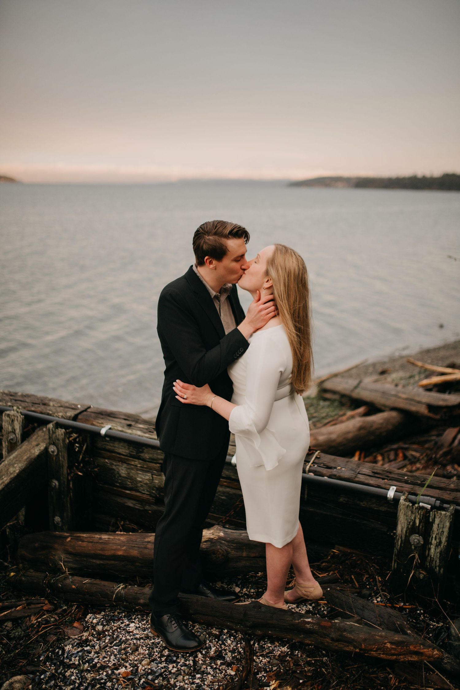 Bride and groom kissing near weathered wooden dock ruins at sunset with serene waterfront backdrop.
