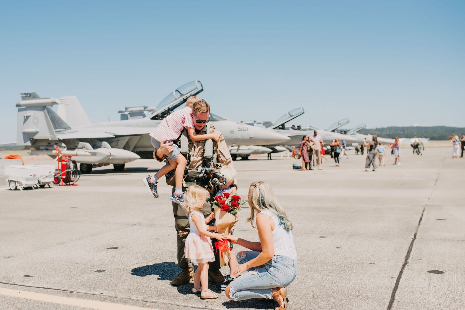 Military father in flight suit lifting child on sunny airfield tarmac with jets in background.