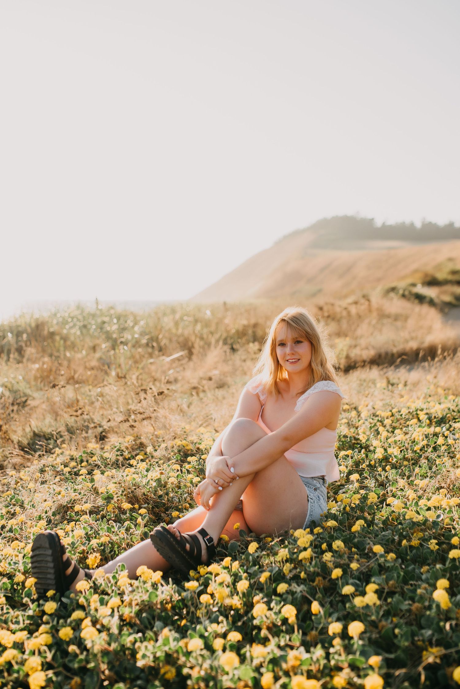 Blonde girl sits among yellow wildflowers on a hillside meadow during golden hour senior photos.