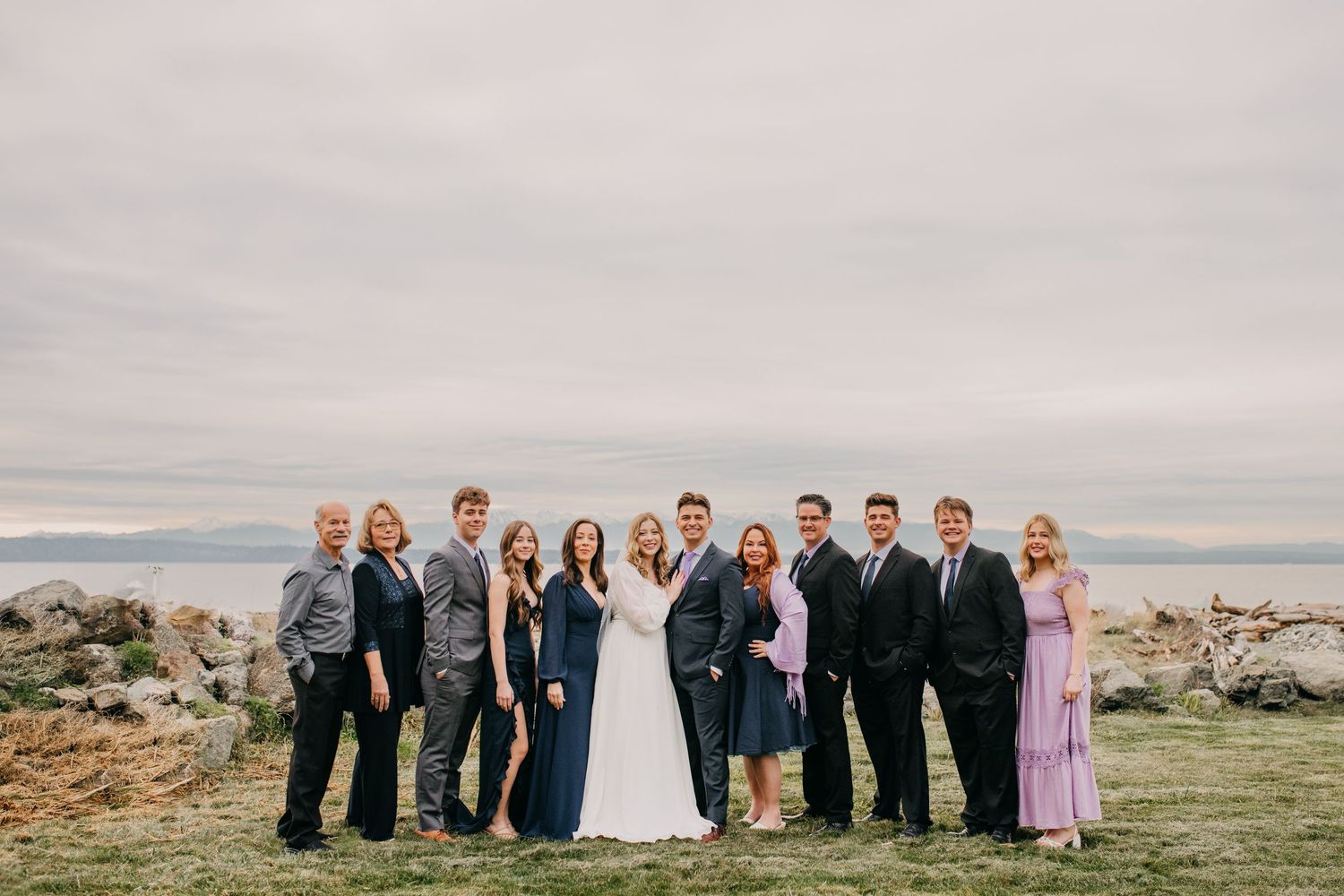 Wedding party group portrait of ten people posing outdoors with overcast sky and water in background.