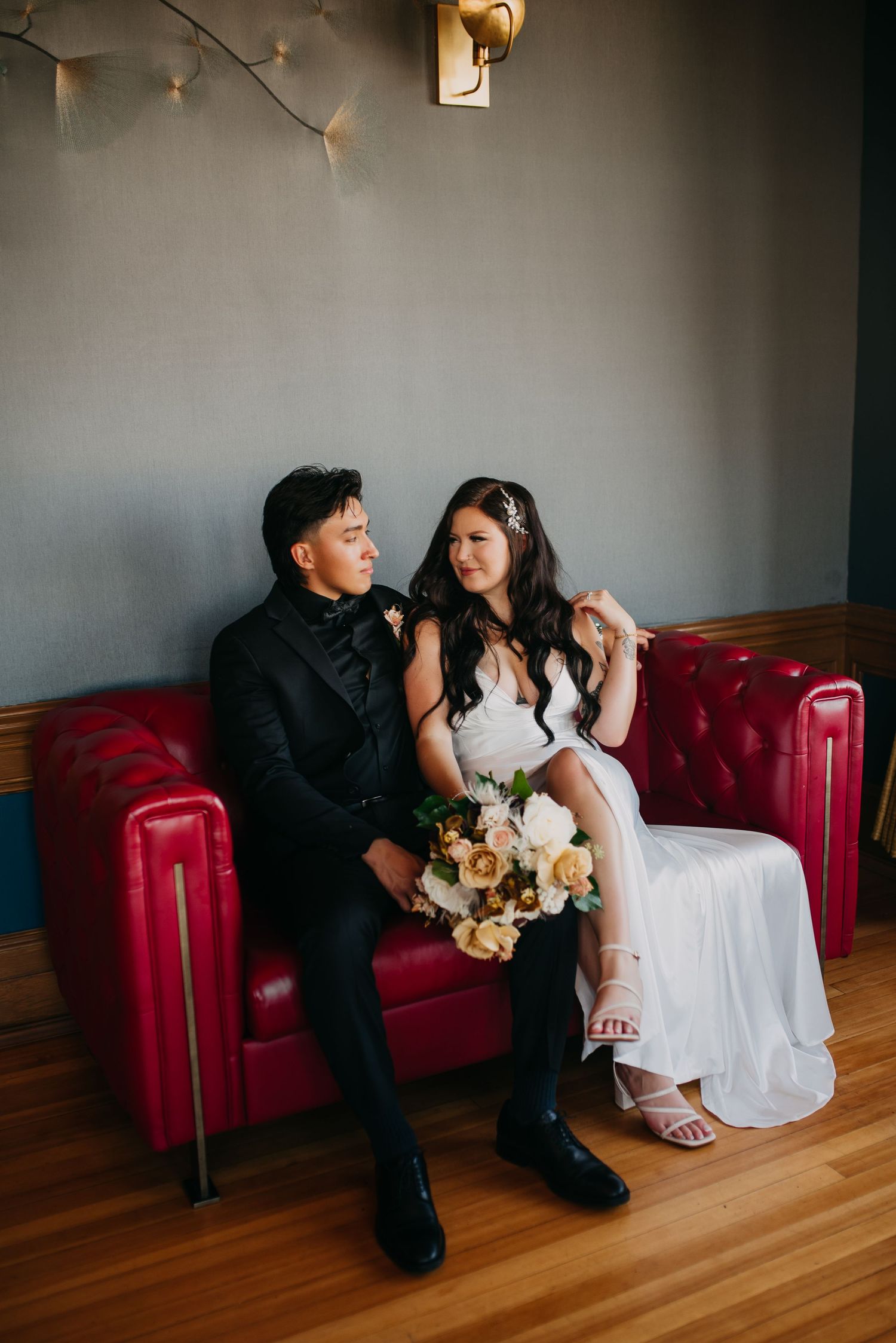 Bride and groom sitting together on a red tufted sofa, bride holding a white and gold bouquet.