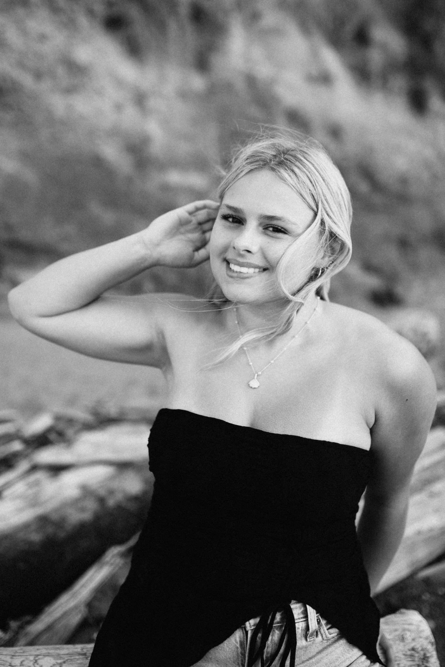 Smiling blonde woman in black strapless top poses on rocky beach in black and white portrait photo.