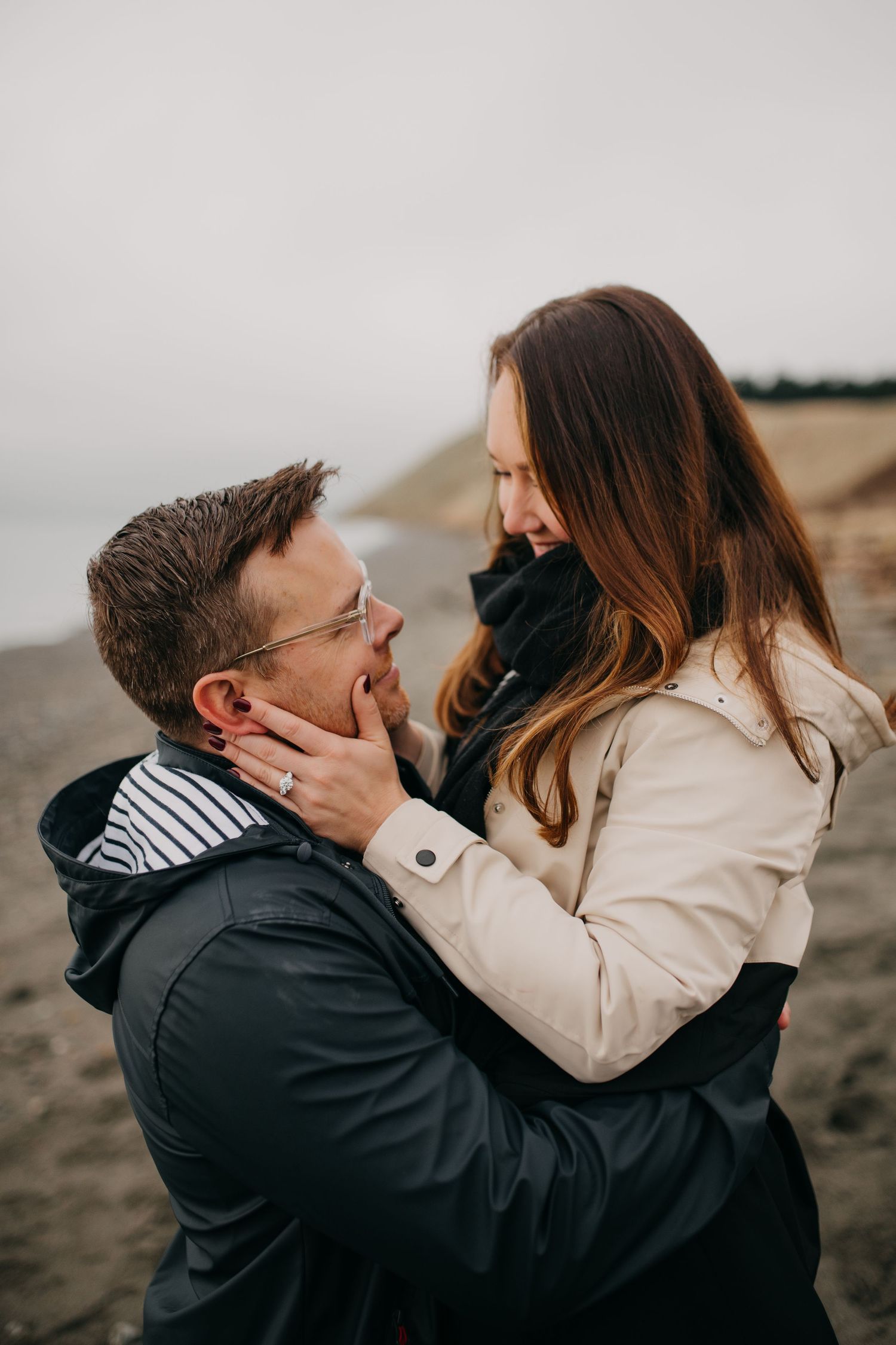 Joyful couple laughing together on beach, woman held by man wearing glasses, engagement photo.