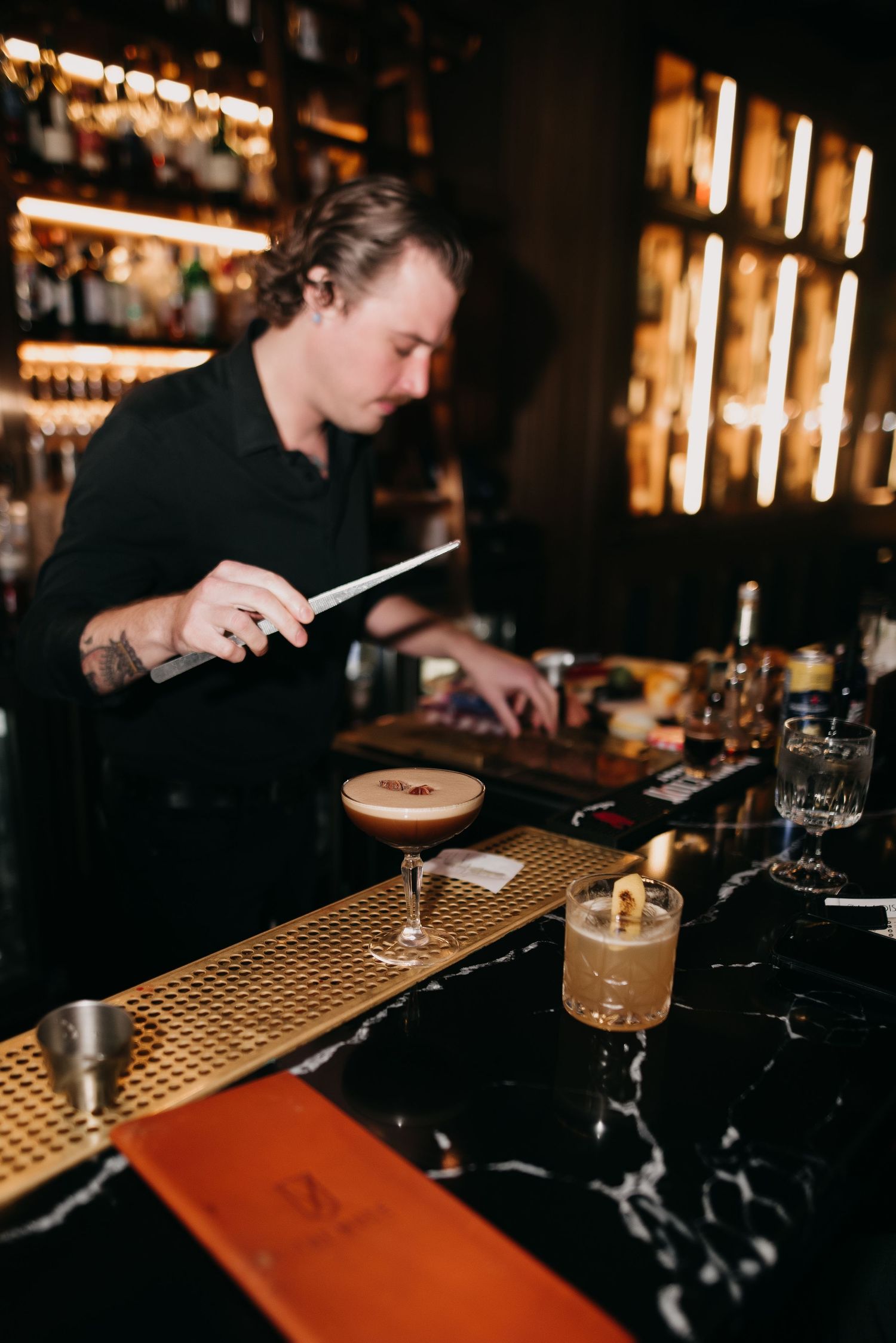 Bartender in black shirt preparing espresso martini cocktails at dark marble bar counter.