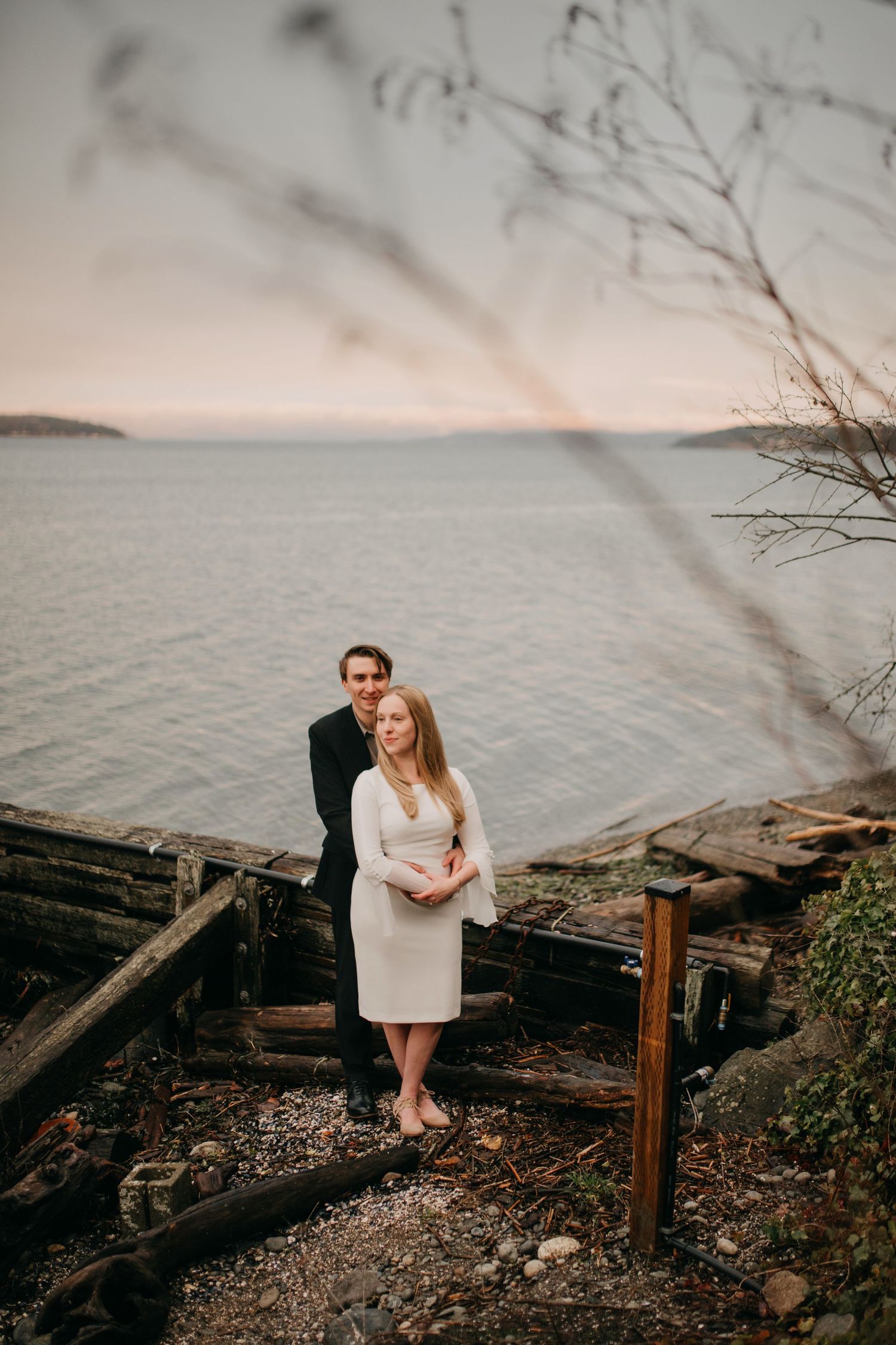 Couple stands together on a weathered dock overlooking a serene bay at dusk during engagement session.
