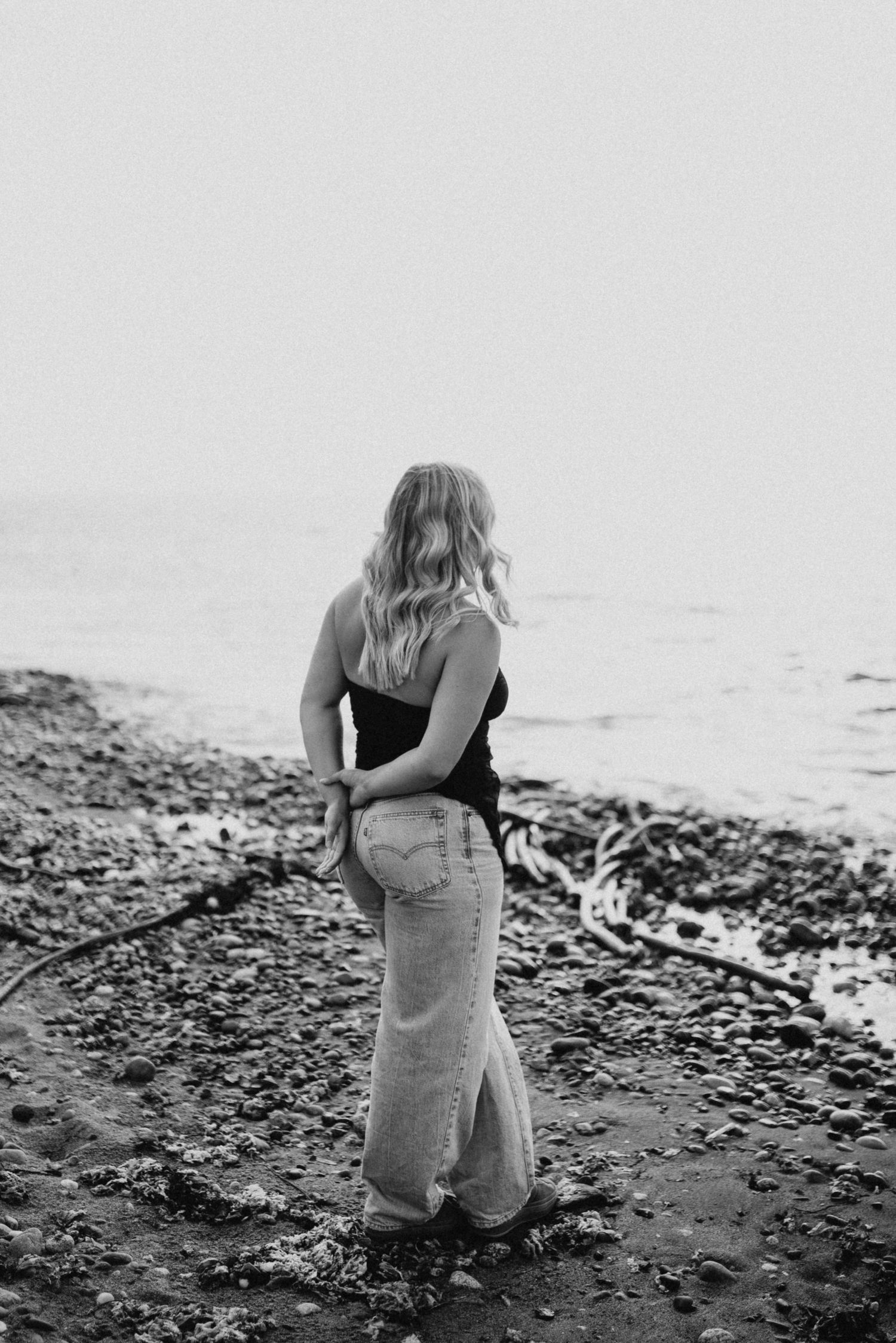 Black and white photo of blonde woman in jeans standing on rocky shoreline facing the water.
