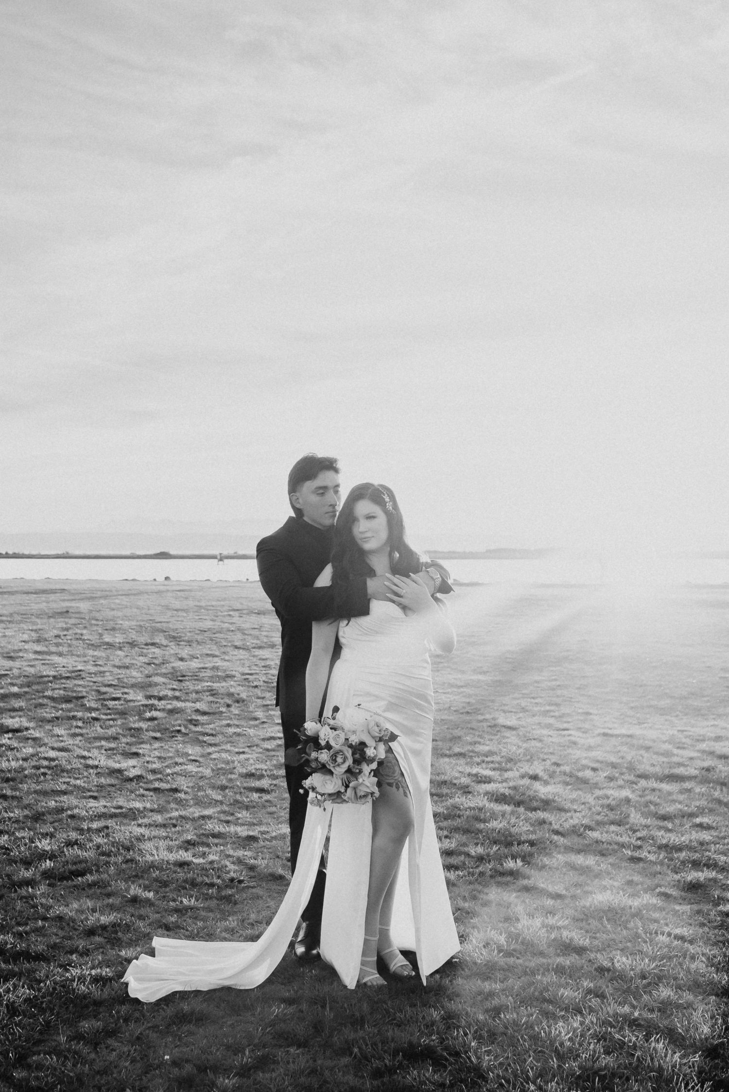 Black and white photo of couple embracing in open field with dramatic sky and bridal train flowing.