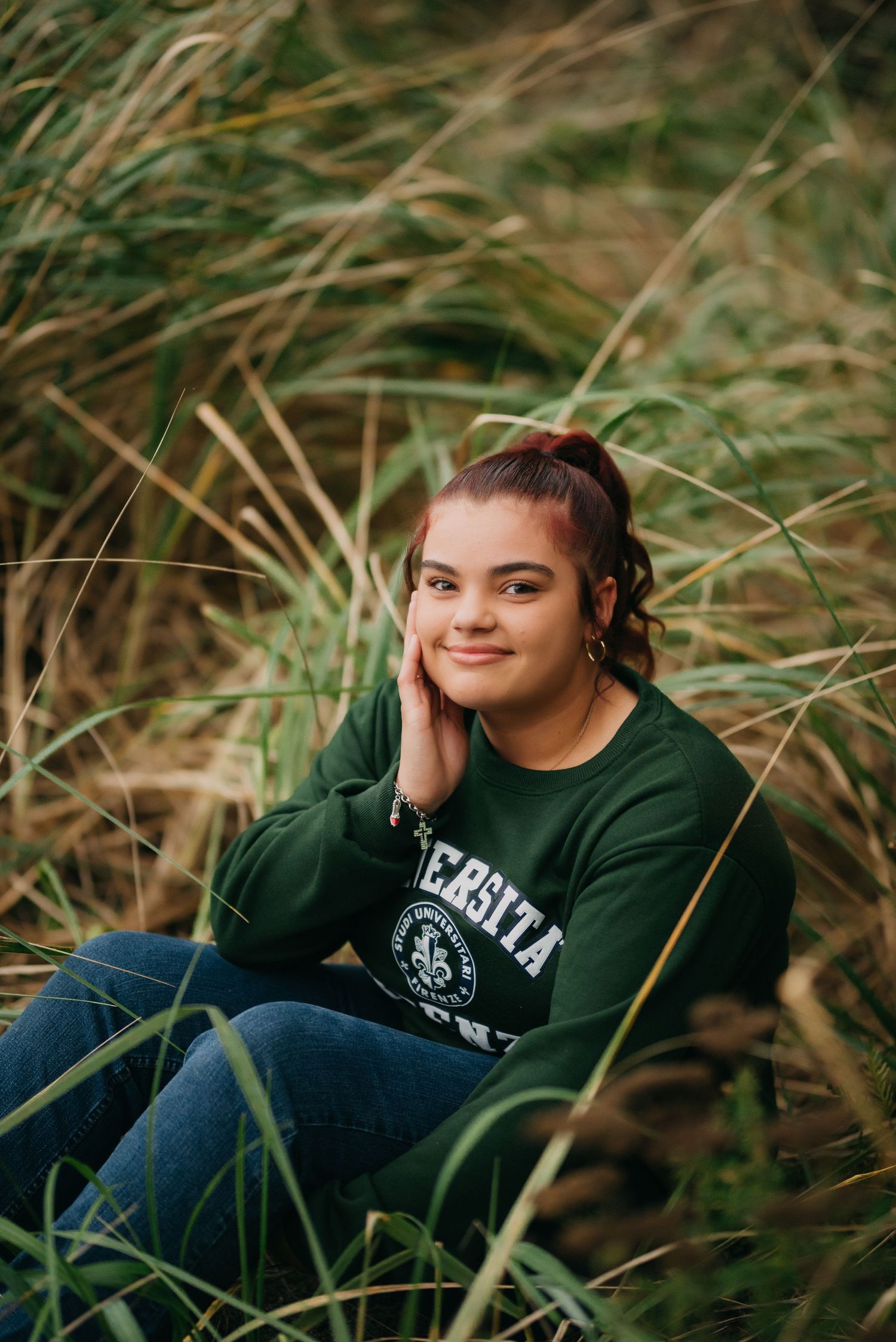 Smiling young woman in green Universita Firenze sweatshirt sits among tall grass with hand on her cheek.