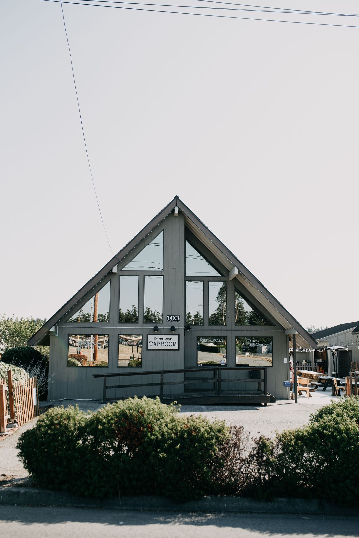 A modern A-frame building with large glass windows serving as a taproom entrance surrounded by shrubs.