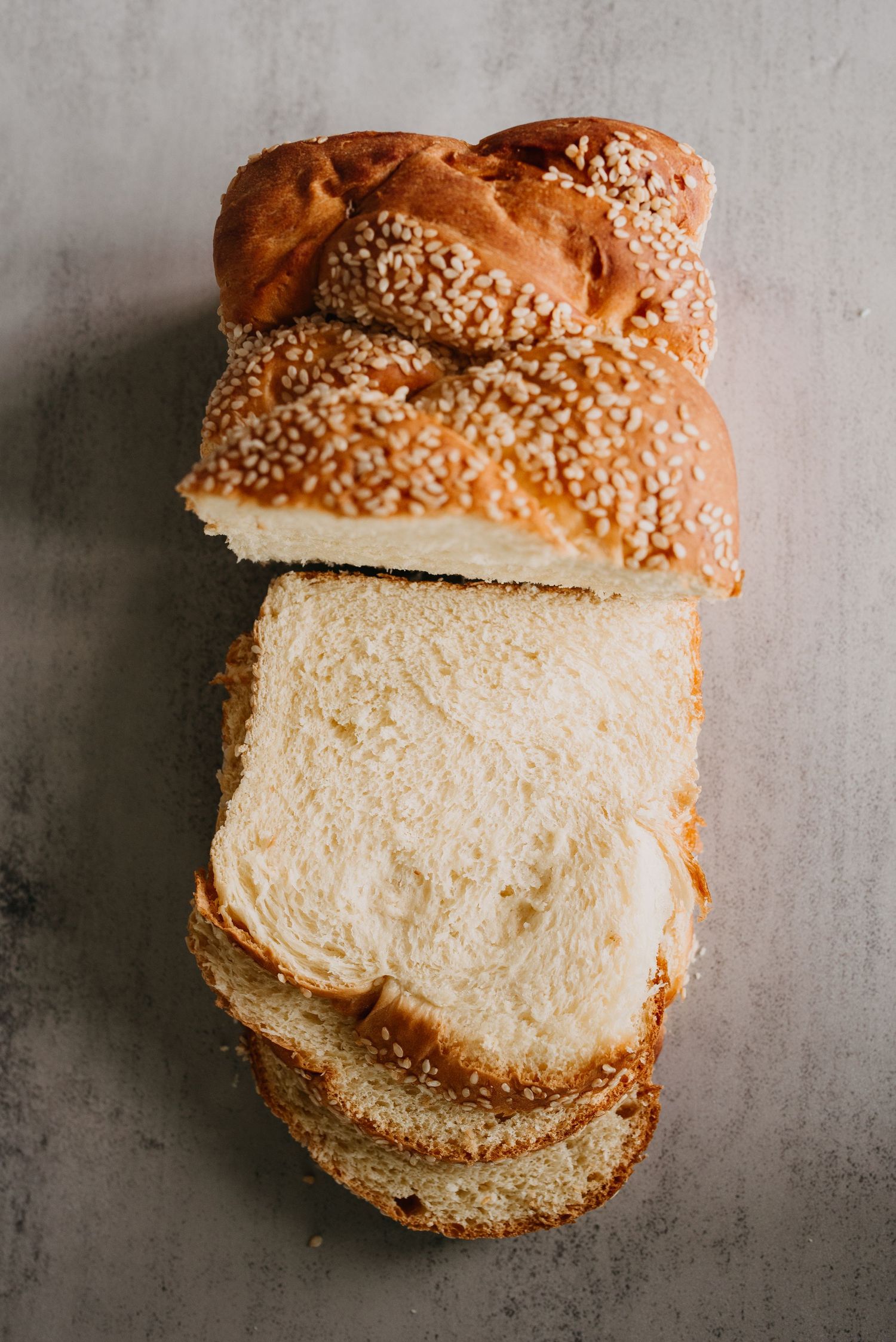 Sliced sesame seed braided bread loaf with soft white interior on gray stone surface.