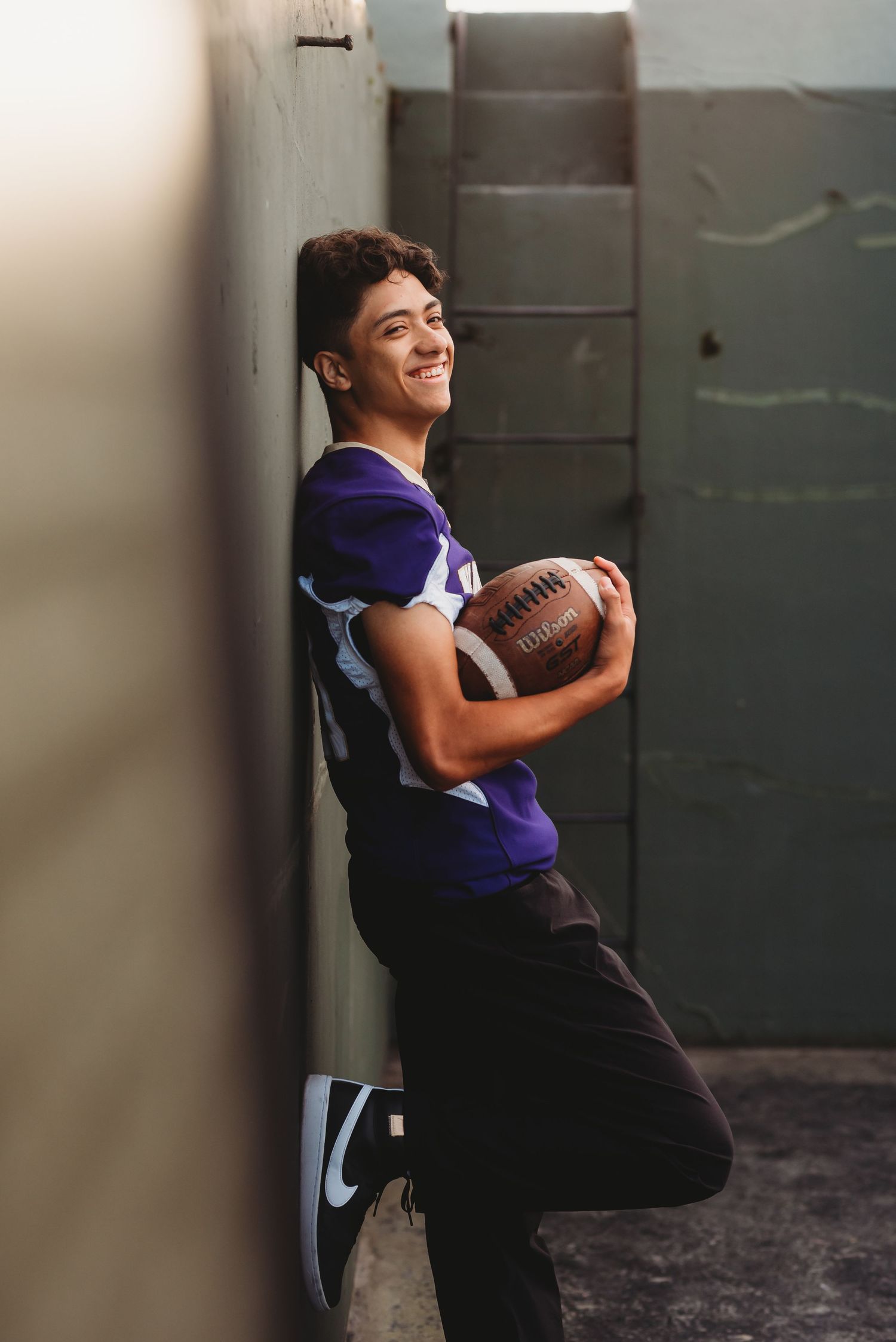 Smiling teenage boy in purple football jersey holding a football while leaning against a wall.