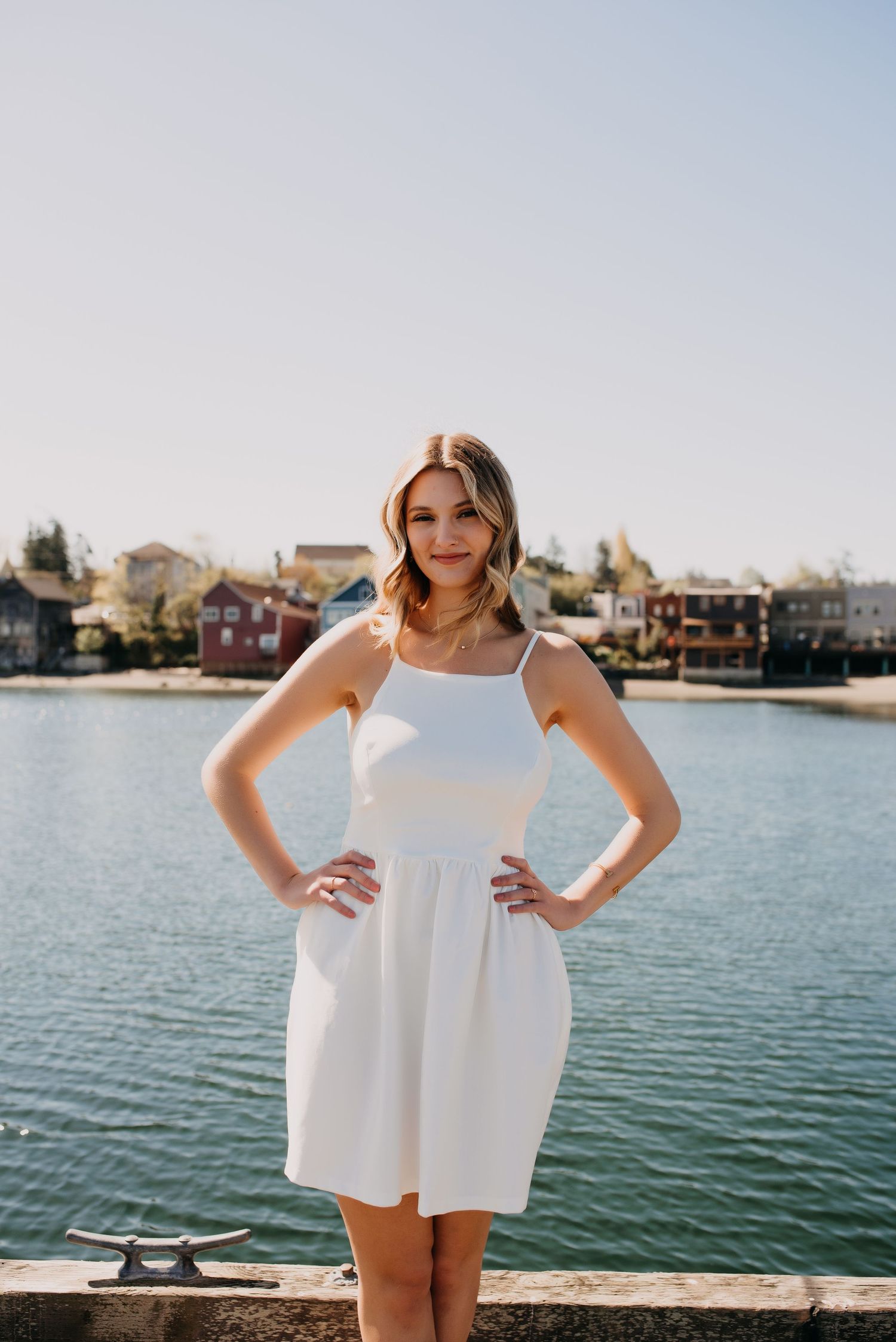 Teen girl in white sundress poses with hands on hips by a scenic waterfront on sunny day.