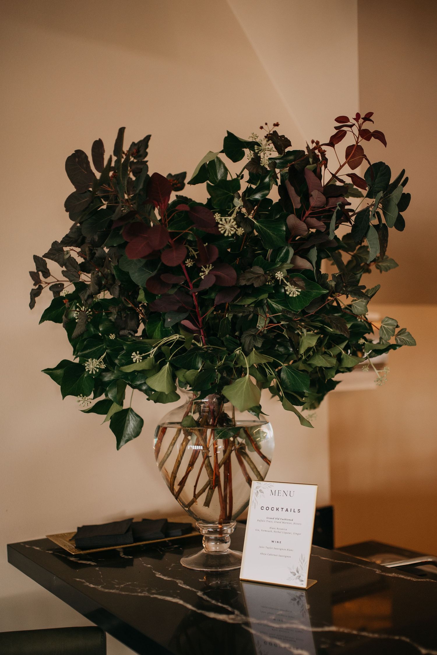 Large lush greenery arrangement with dark foliage in a glass vase on a black marble table.