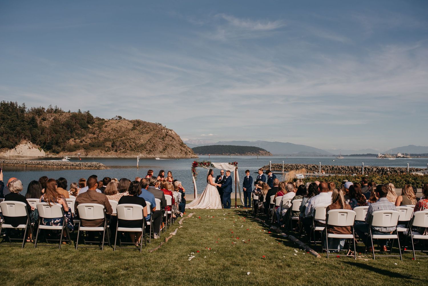 Outdoor waterfront wedding ceremony with guests seated in white chairs overlooking scenic rocky coastline.