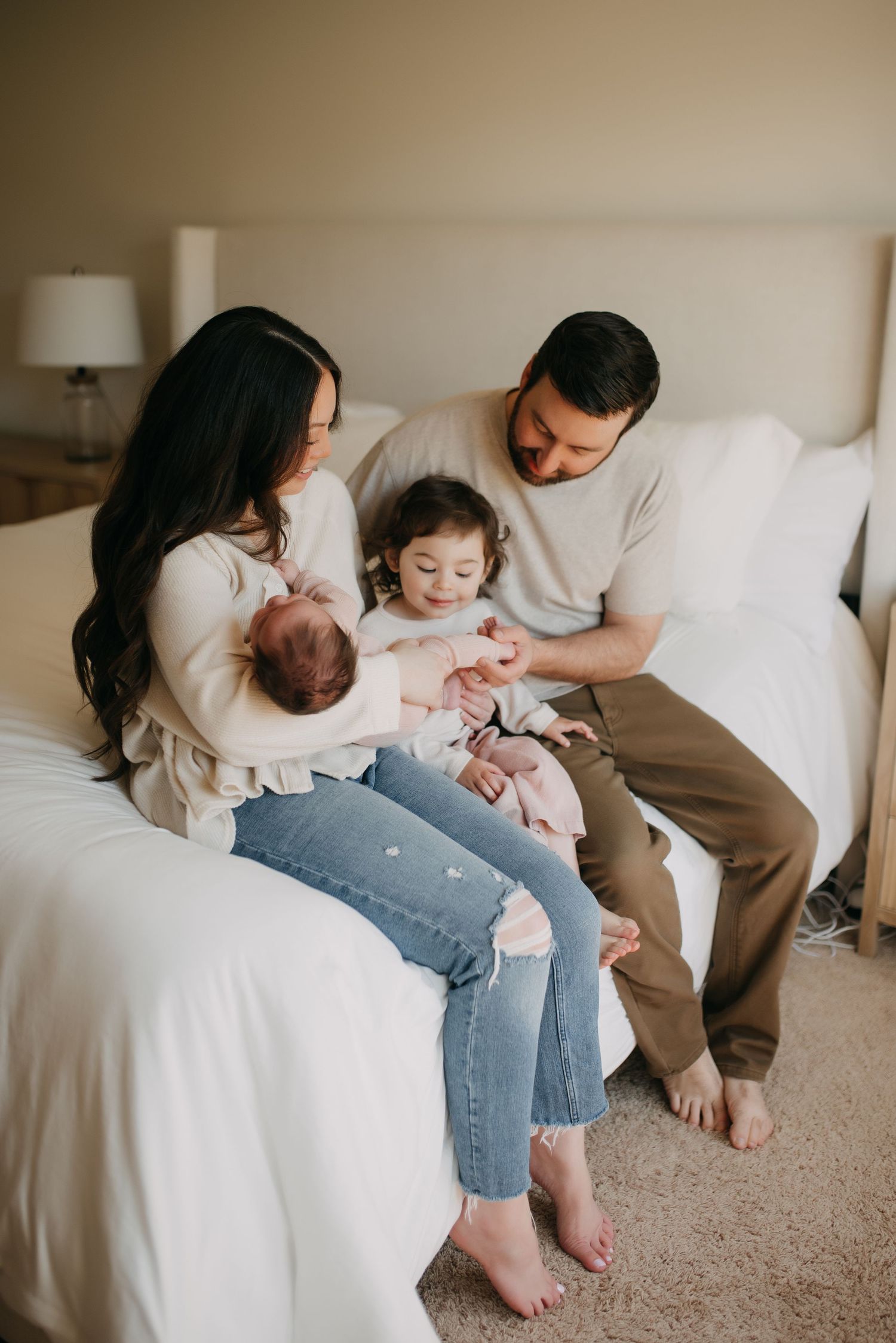 Family of four sitting on bed, mother holding newborn while father and toddler interact warmly.