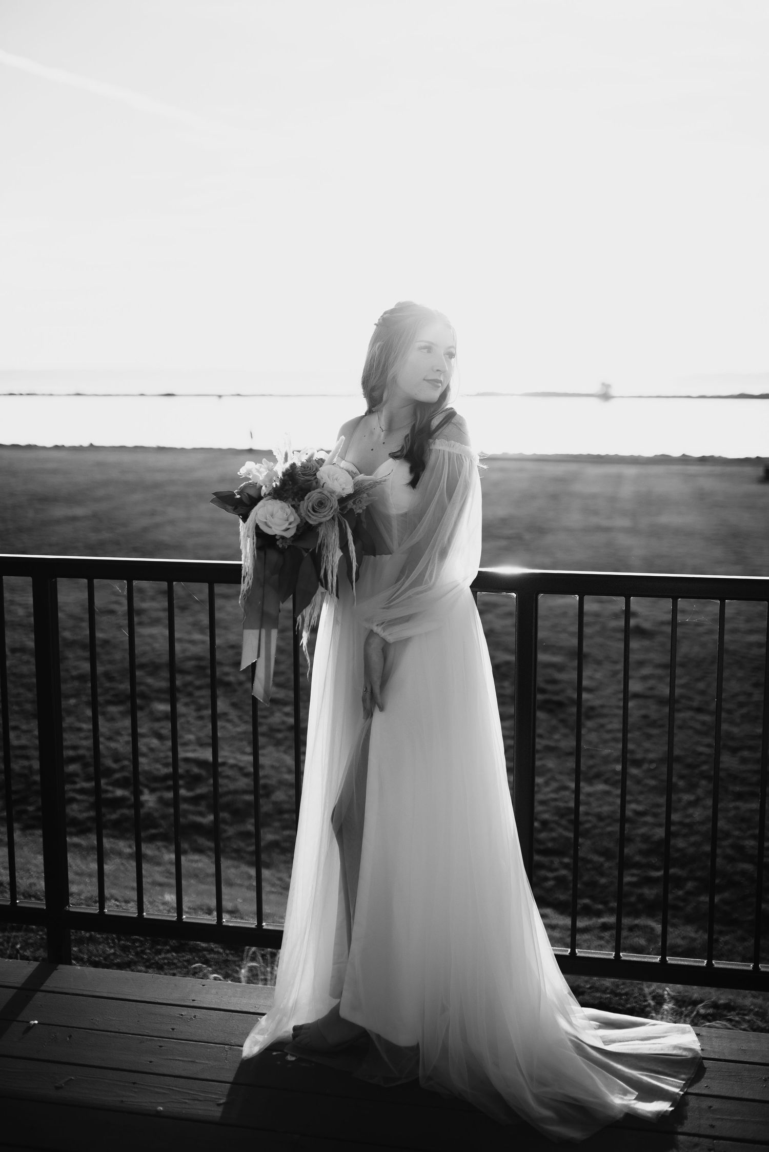 Black and white portrait of a bride in a flowing gown holding a bouquet on a sunlit deck.
