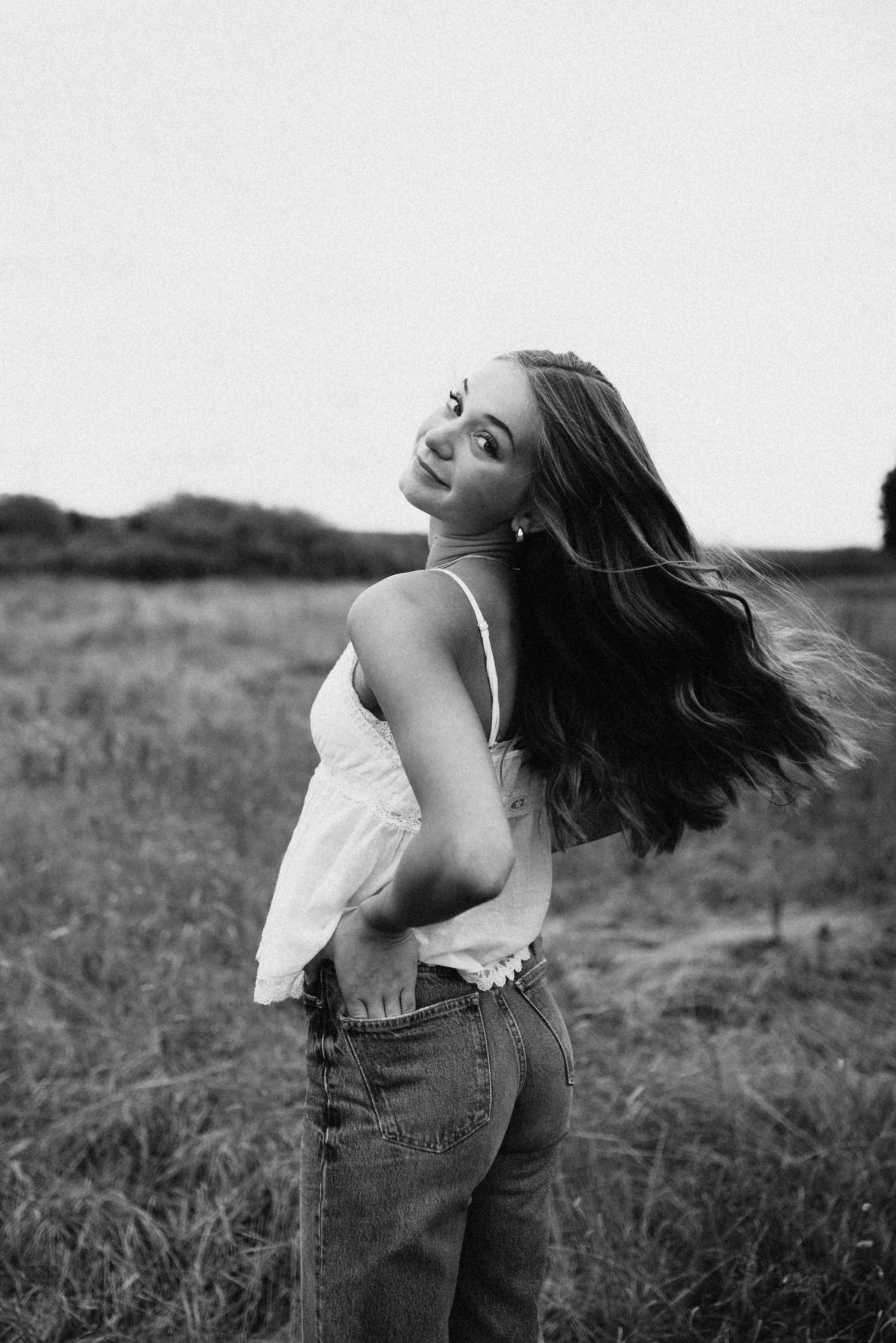 Young woman with flowing dark hair poses in field wearing white camisole and jeans in black and white.