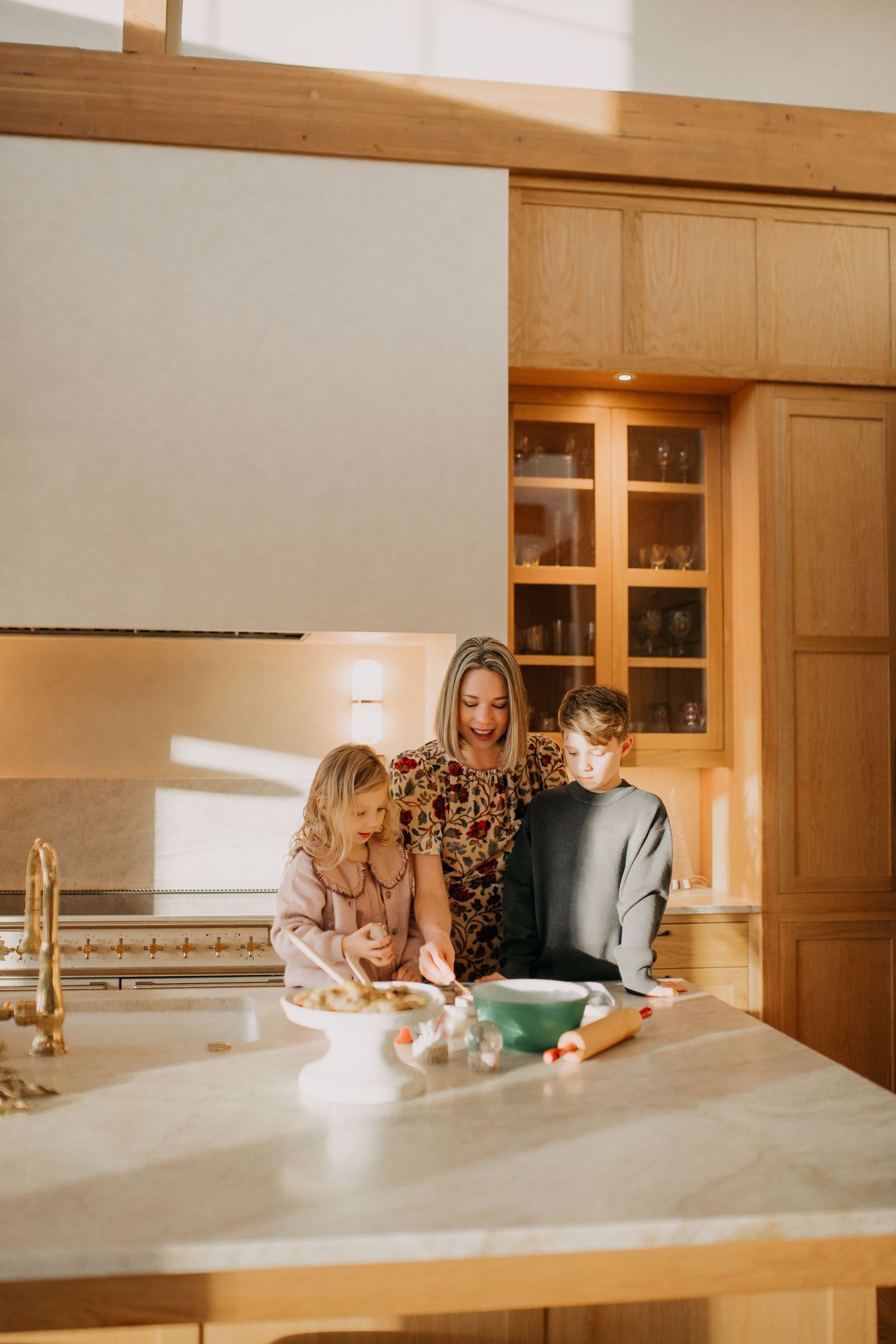 Mother and two children baking together in a bright, modern kitchen with warm wood cabinetry.